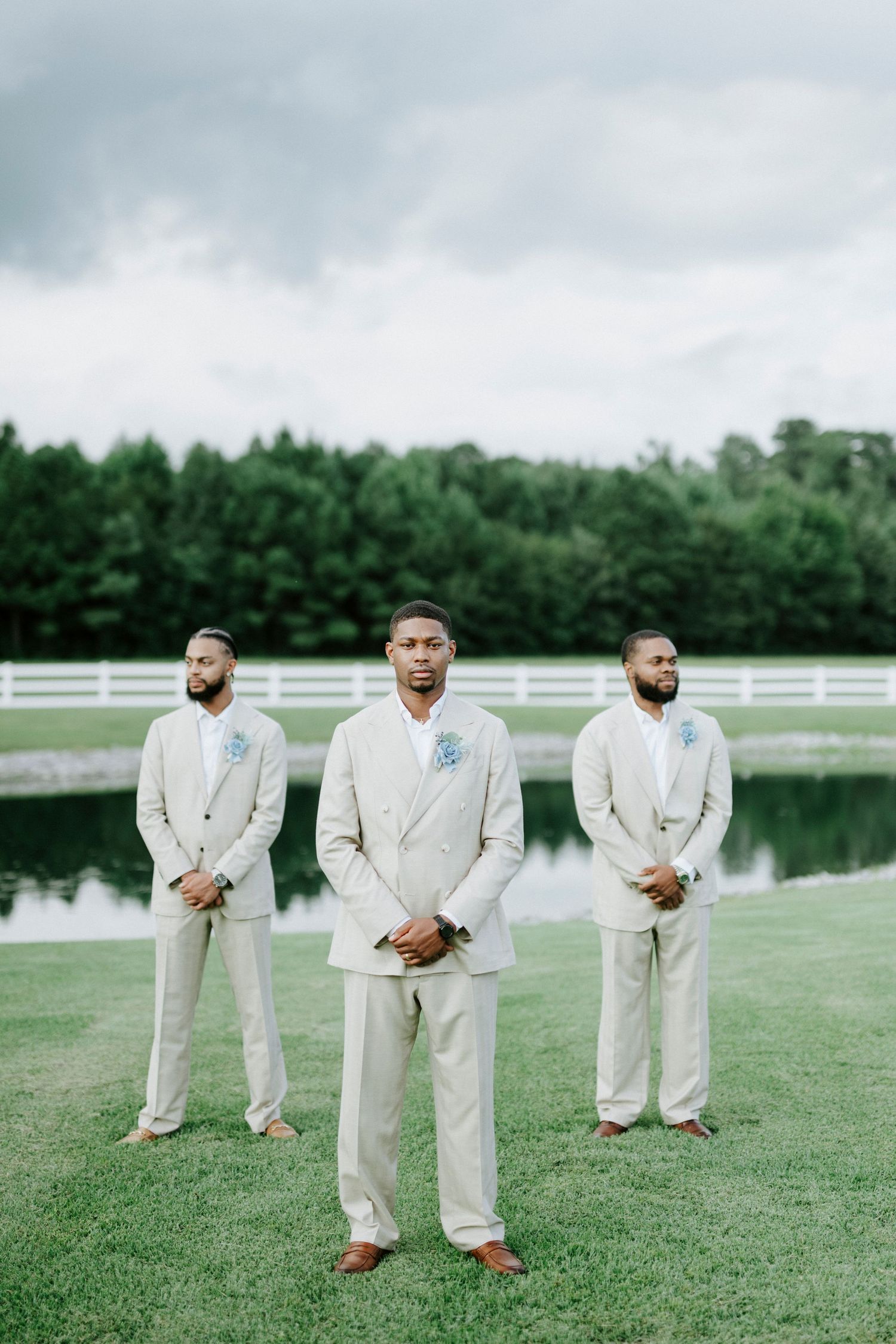 Wedding party in cream suits stand formally on manicured lawn with pond and tree line in background.