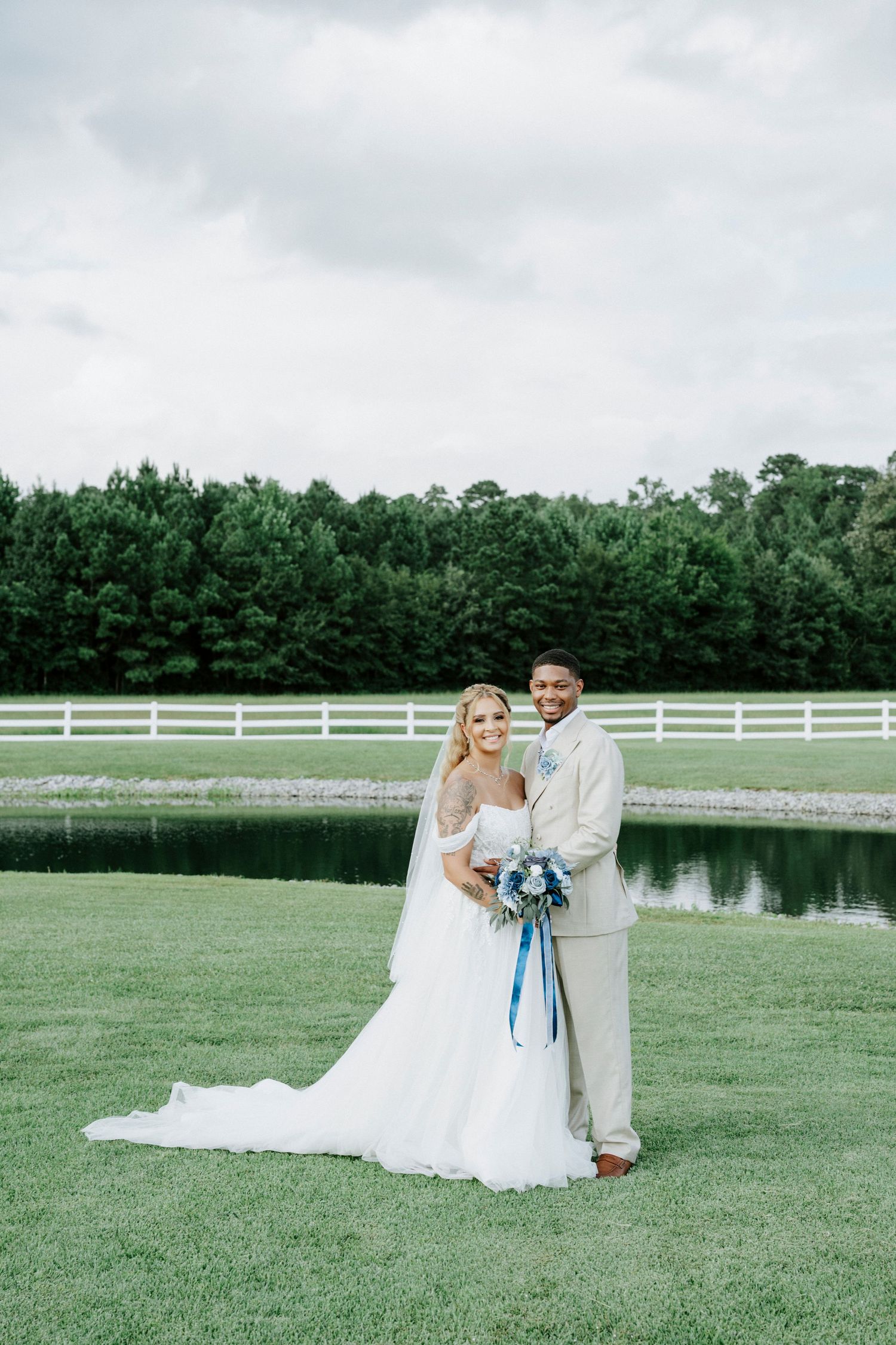 Newlyweds pose together on grass with blue bouquet against white fence and pond backdrop.