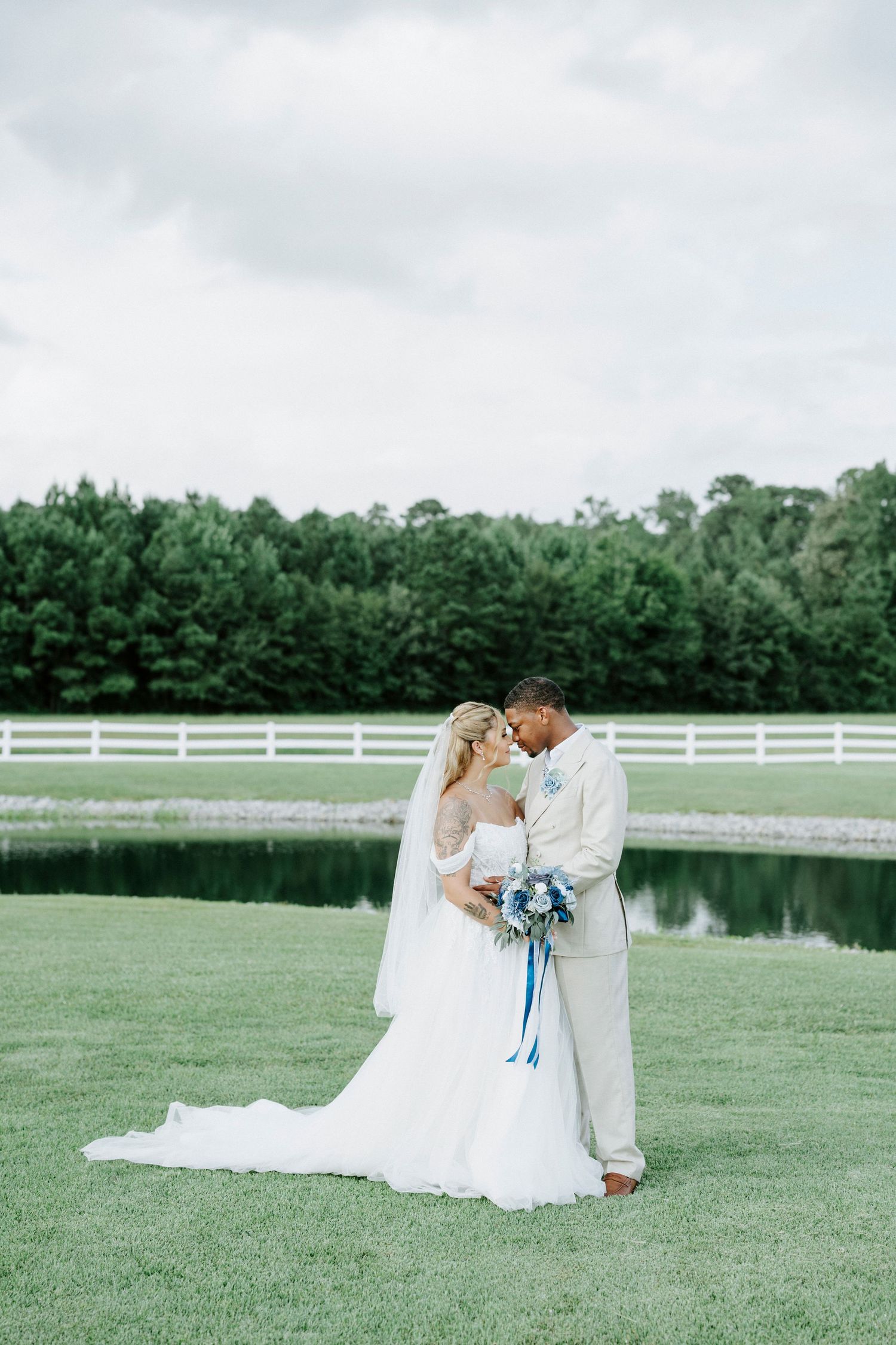 Romantic wedding portrait of couple embracing on lawn with pond and white fence in background.