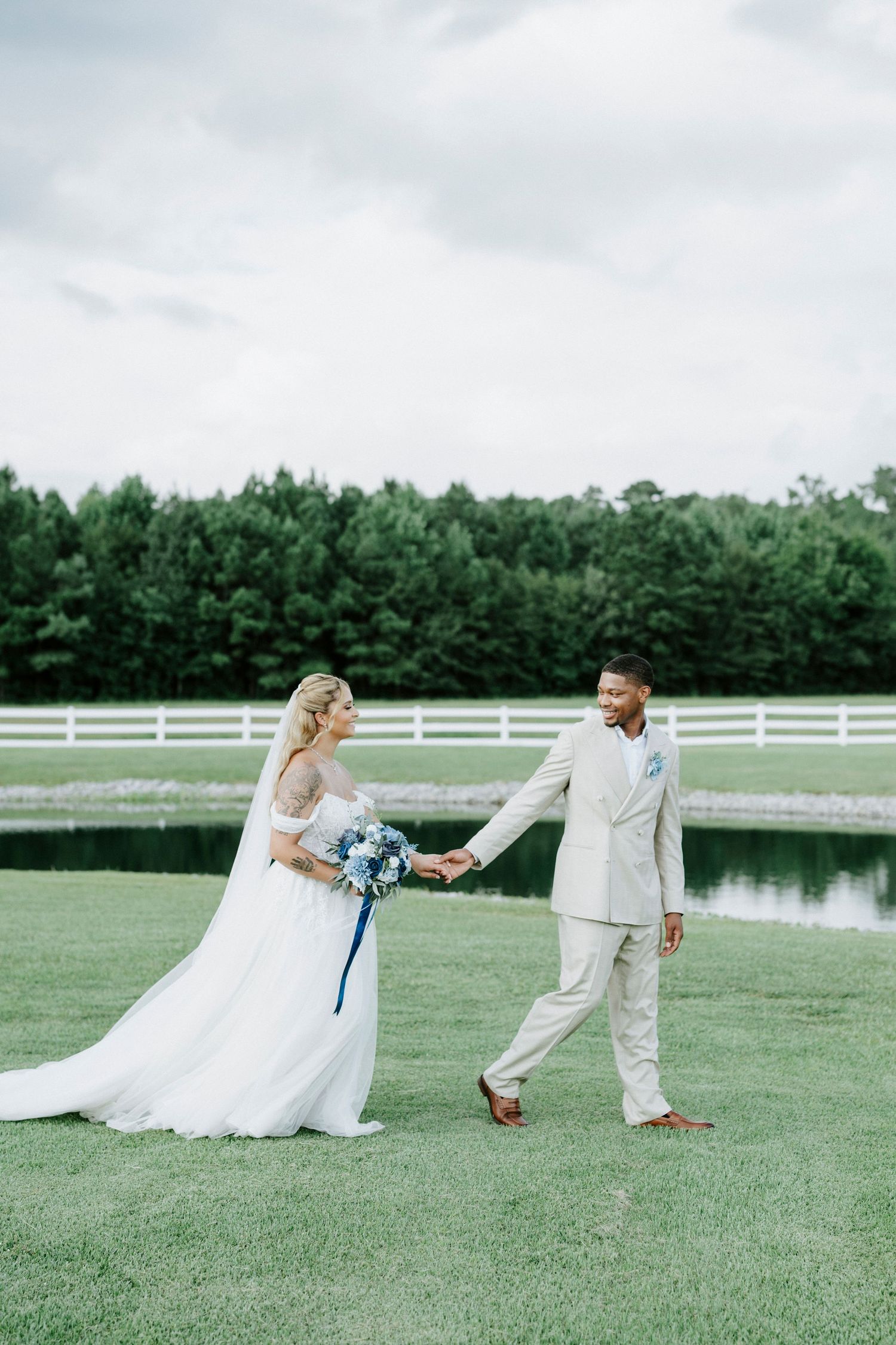 Couple in wedding attire stroll hand in hand on green lawn with blue bouquet and scenic backdrop.