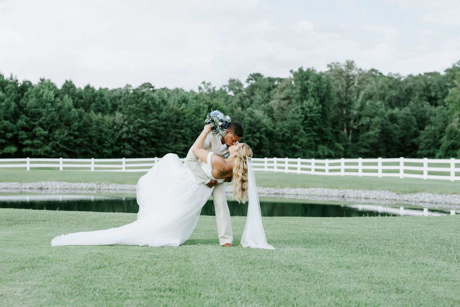 Romantic dip pose of newlyweds on manicured lawn with pond and white fence background.