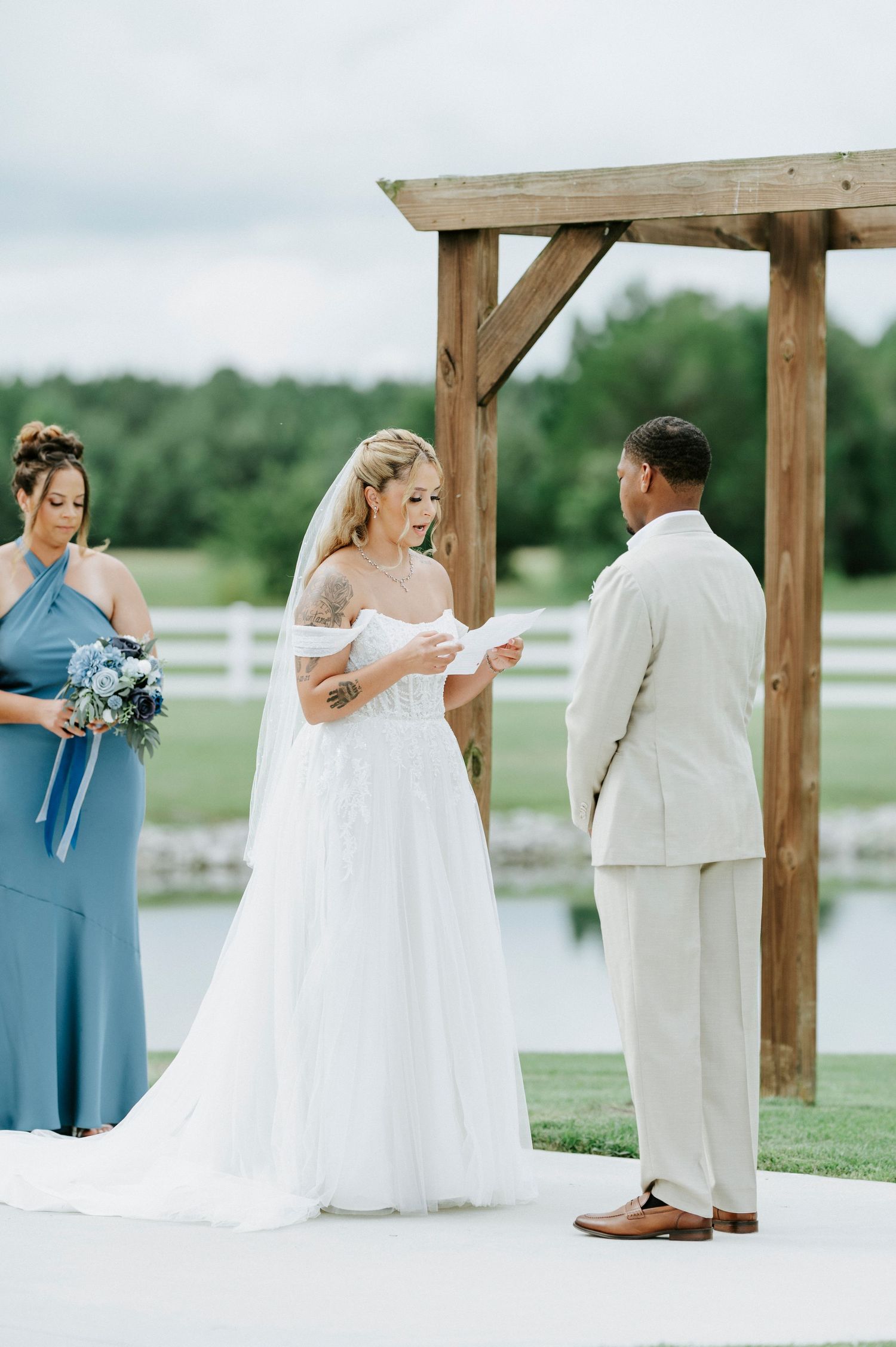 Wedding party gathered under a wooden pergola by a lake during an outdoor ceremony.