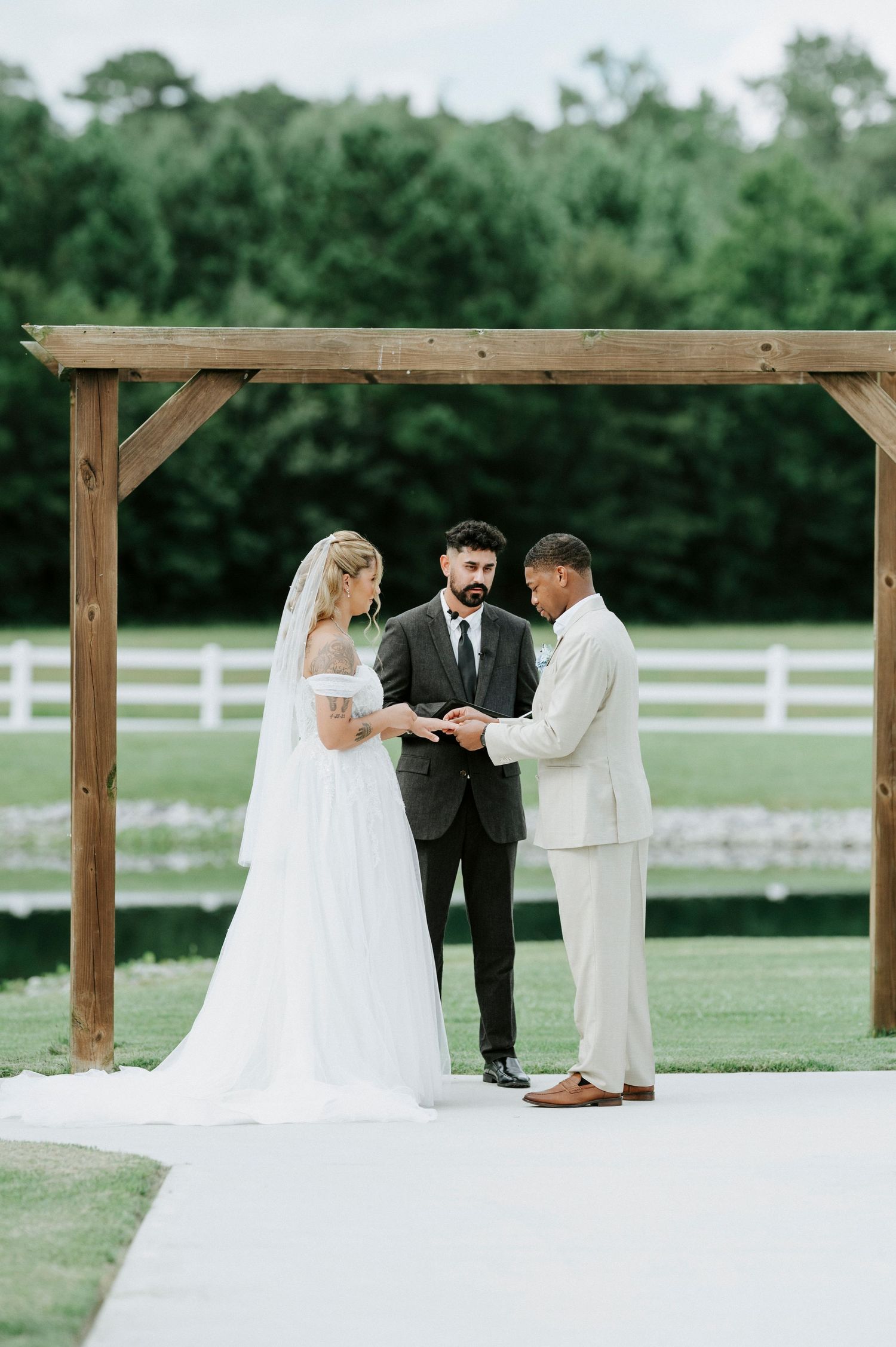 Wedding ceremony taking place under a rustic wooden arbor with white fencing in the background.