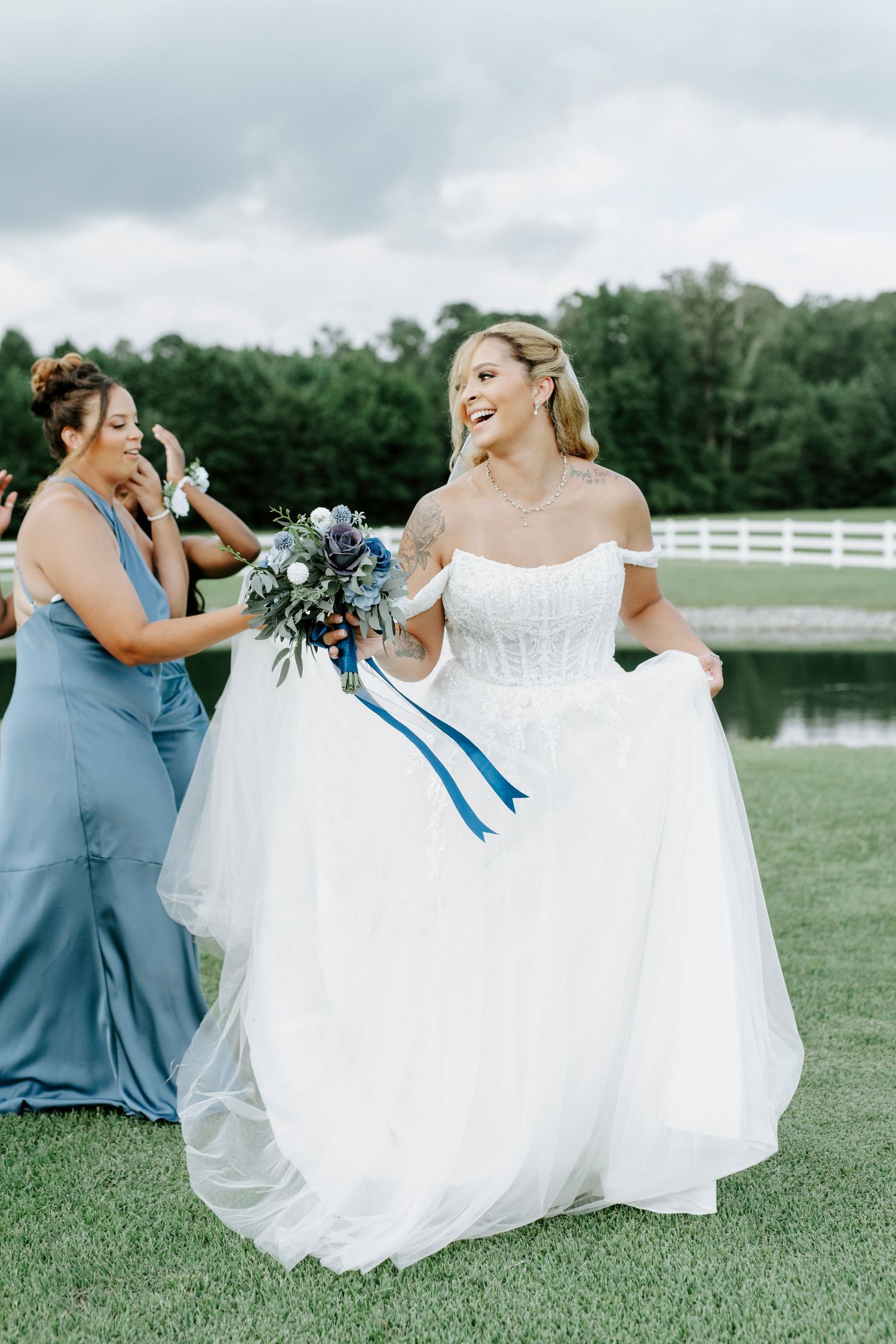 Wedding party in blue dresses helping arrange the bride's flowing white gown on grass.