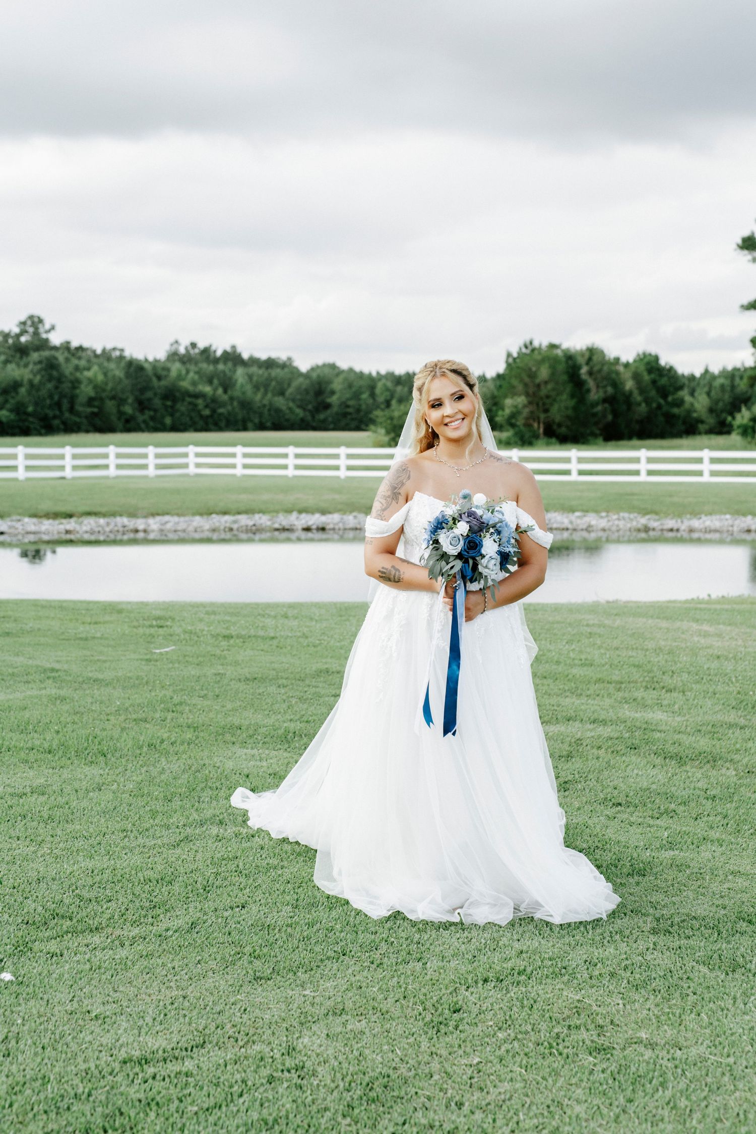 Bride in white gown holding blue bouquet poses in front of white fence on grassy field.