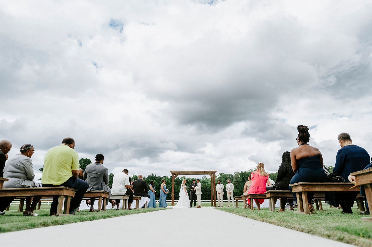 Wide shot of wedding guests seated outdoors under cloudy skies.