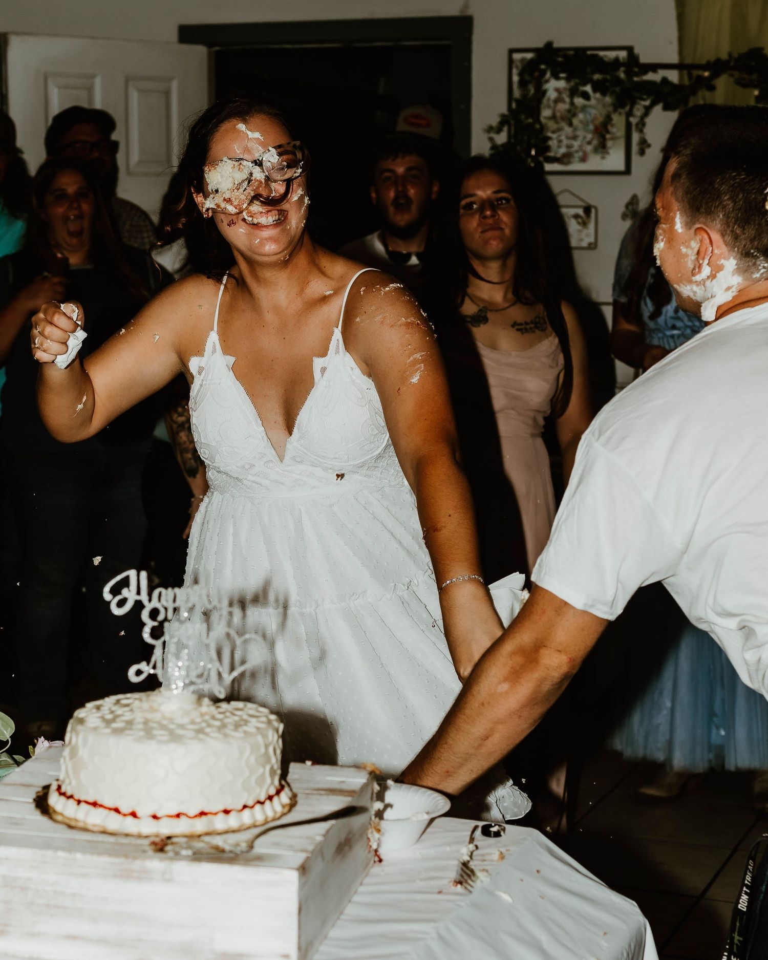 Wedding reception scene with white cake and guests gathered around a table.