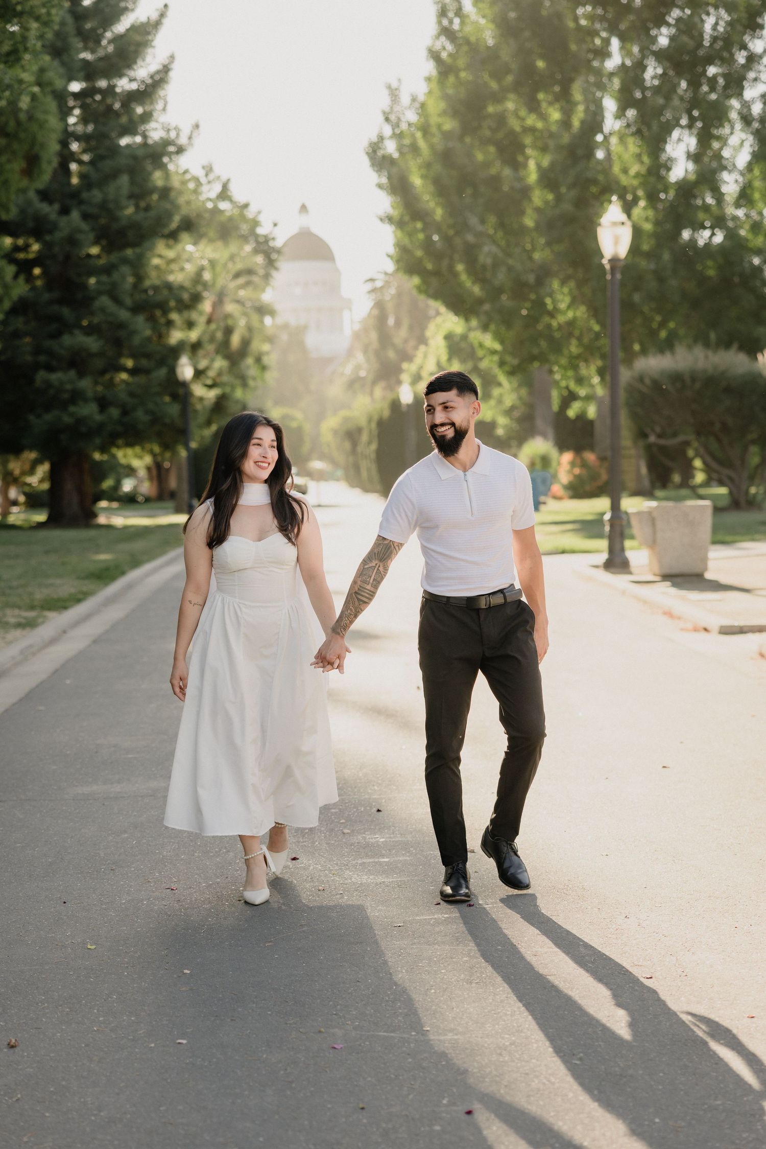 A couple holding hands walks down a tree-lined path on a sunny day, with the bride in white and groom in casual attire.