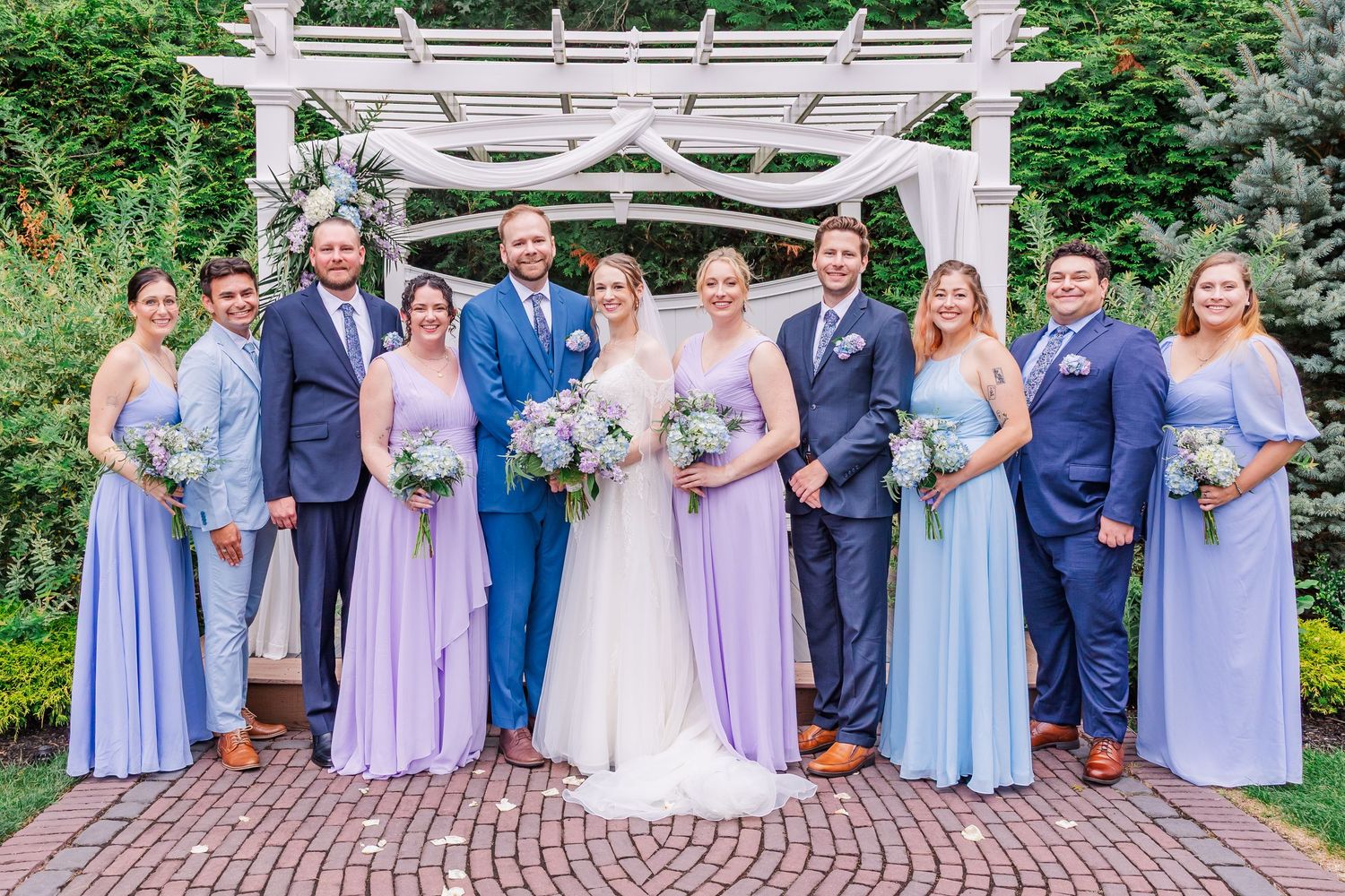 Wedding party wearing coordinated navy and lavender attire poses together under white garden arbor on brick patio.