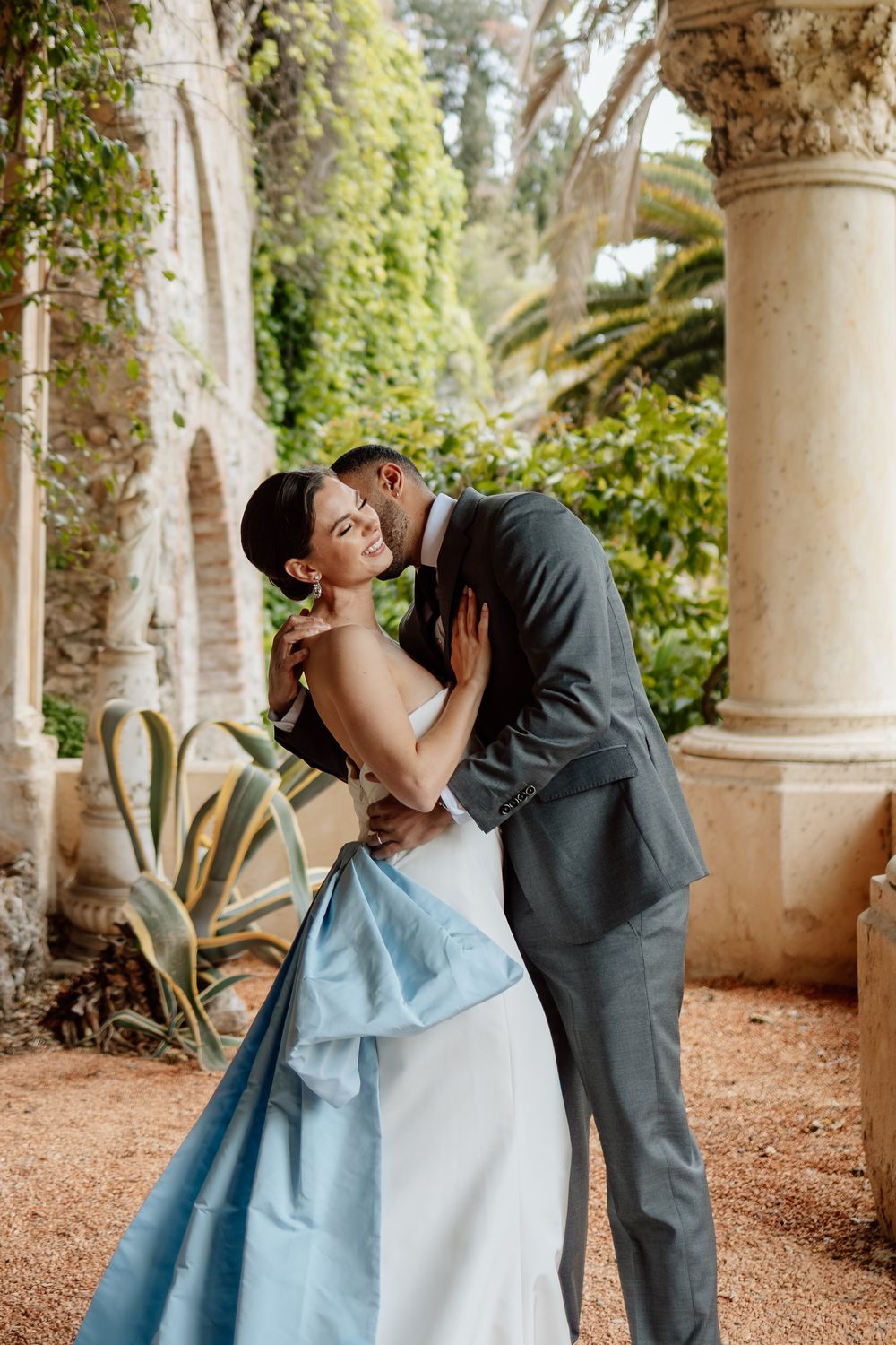 Two elegantly dressed figures share a romantic moment beneath stone pillars in a garden setting. Italian destination wedding photographer.
