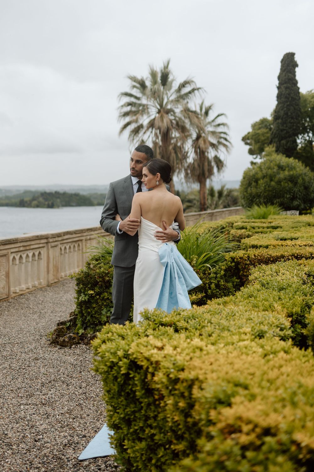 A couple shares an intimate moment on a stone pathway overlooking a scenic Mediterranean coast with lush greenery. Destination wedding photo