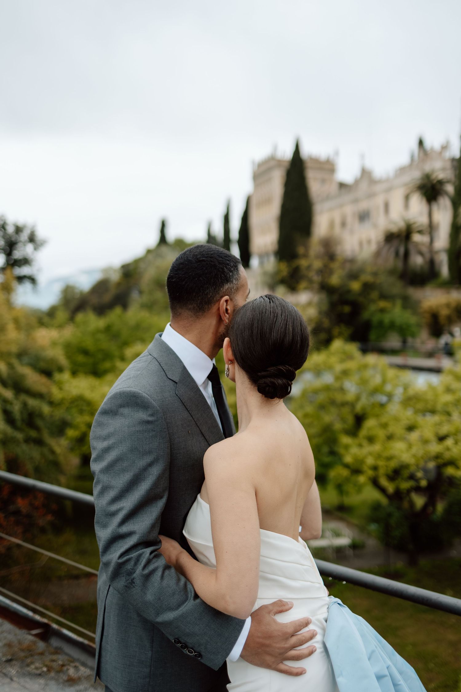 A couple embraces while looking at a beautiful Mediterranean villa with cypress trees and palm trees in the background.