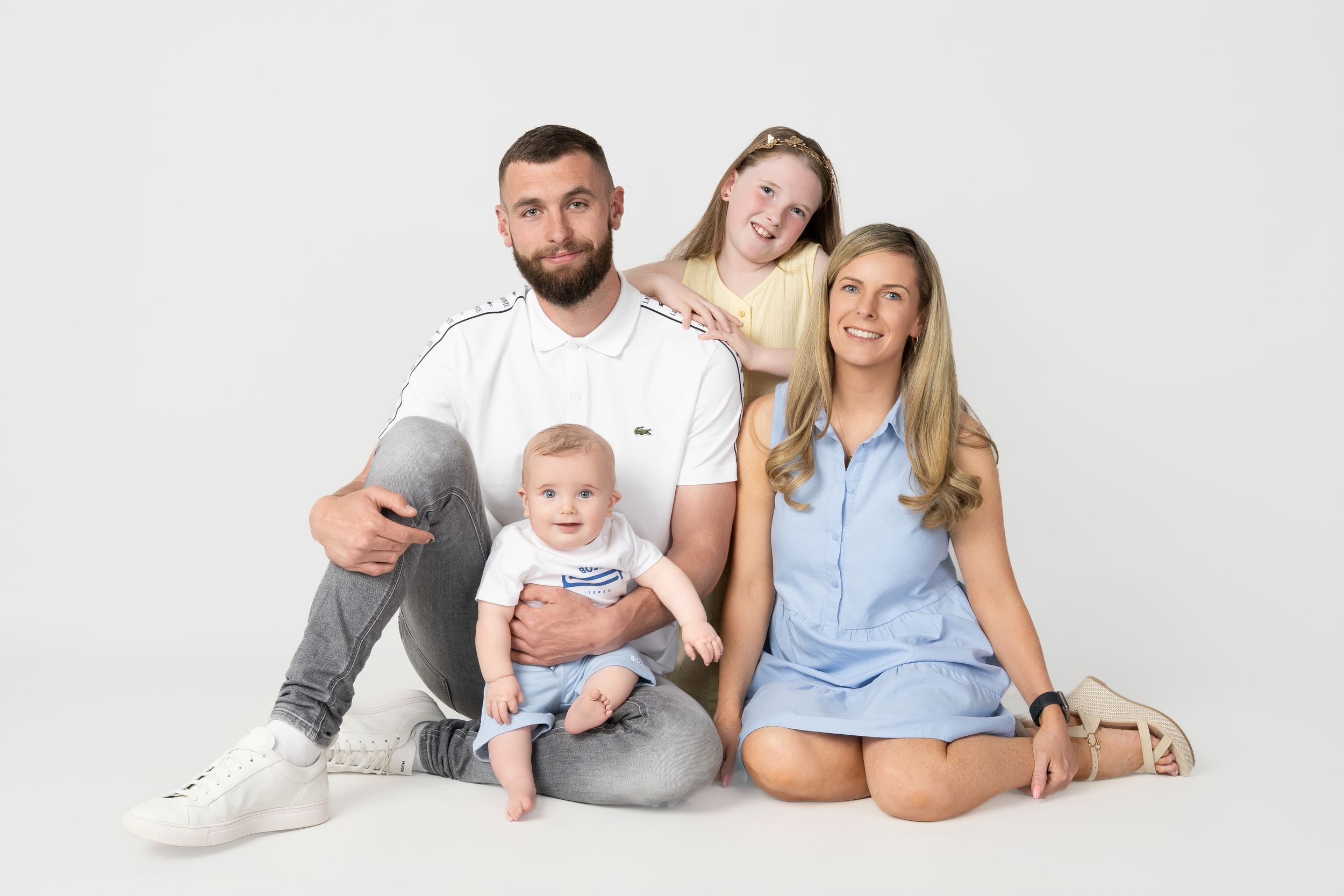 A family group poses together on white background wearing casual clothing in soft neutral colors.