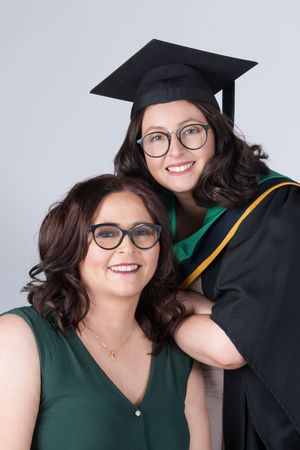 A graduation photo featuring two smiling individuals wearing glasses with one in academic regalia and cap against a light background.