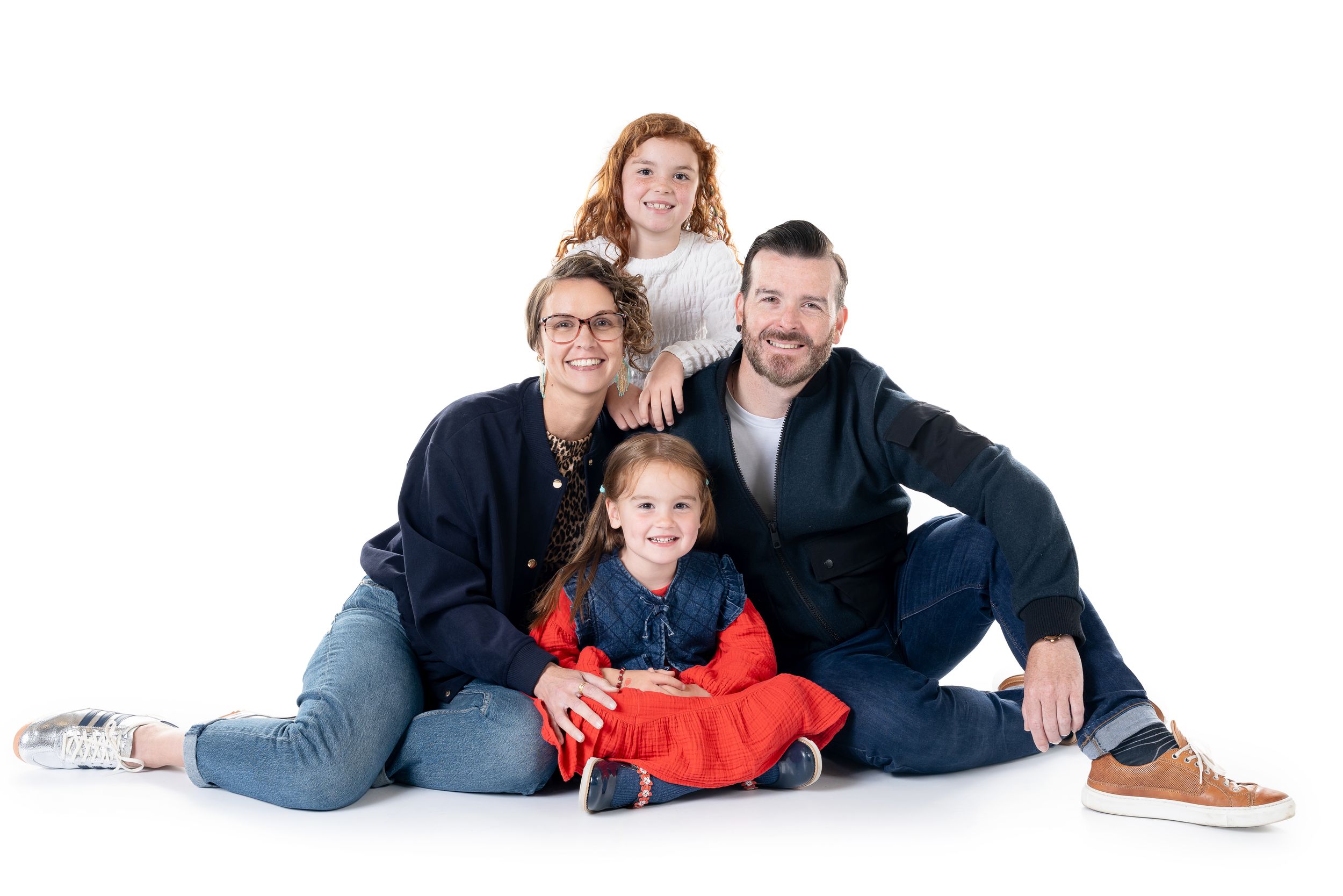 A family of four poses together on a white studio backdrop wearing casual clothing in blue and red tones.