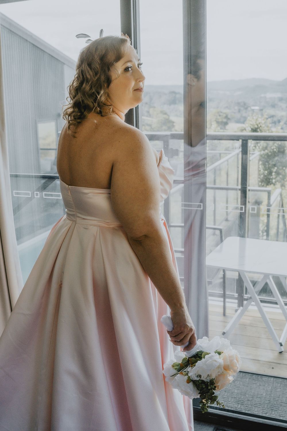 A person in a blush pink strapless gown holds a white floral bouquet while gazing out a window with scenic views.