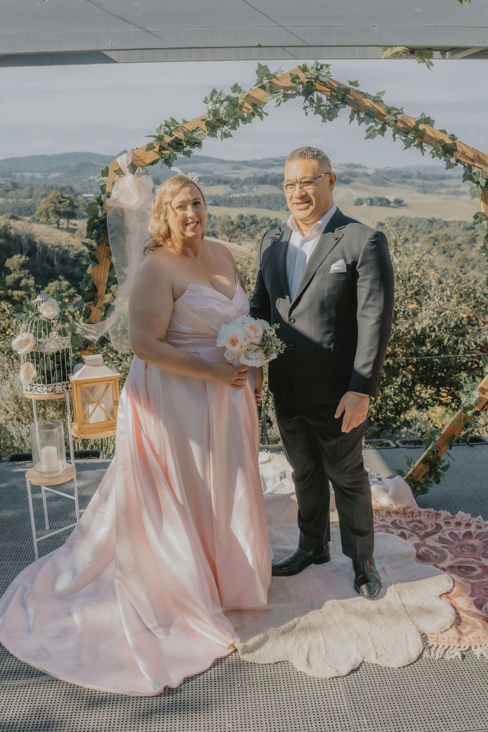 A bride in a blush pink gown and groom in a dark suit stand under a floral arch on a hilltop wedding venue.