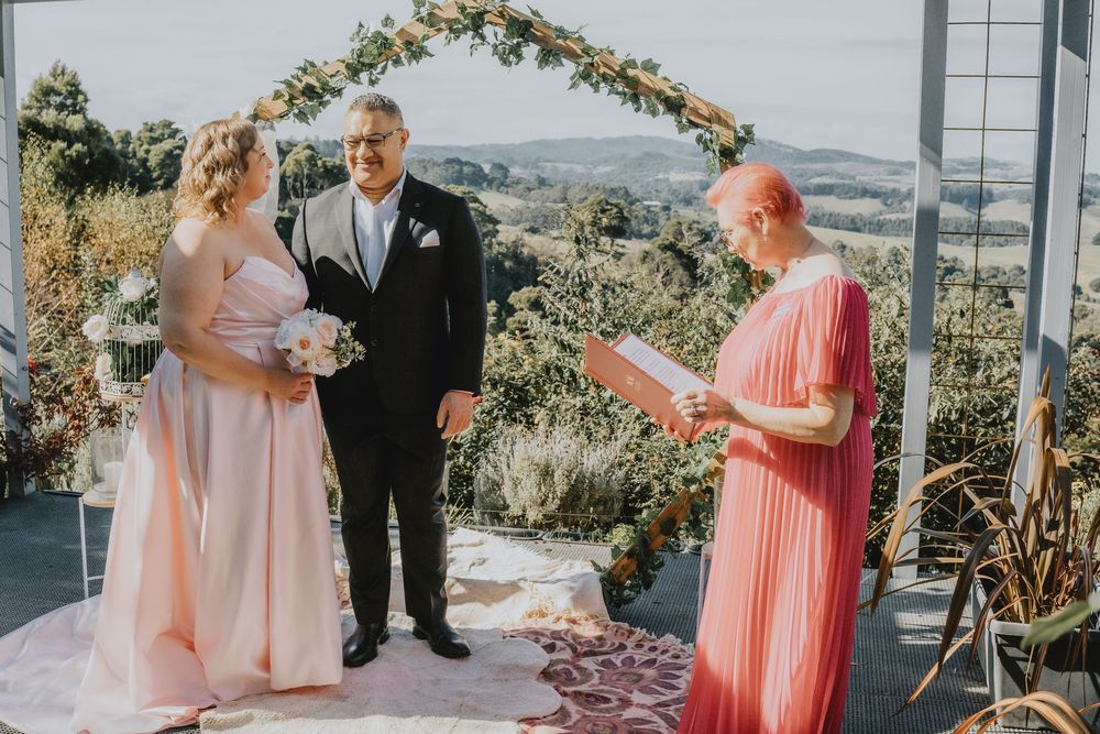 A wedding ceremony takes place under a floral arch with mountain views in the background.