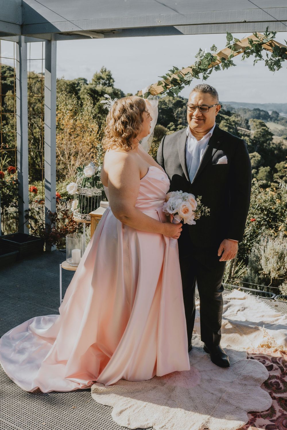 A romantic outdoor wedding ceremony with a couple in blush pink gown and black suit on a scenic hilltop veranda.