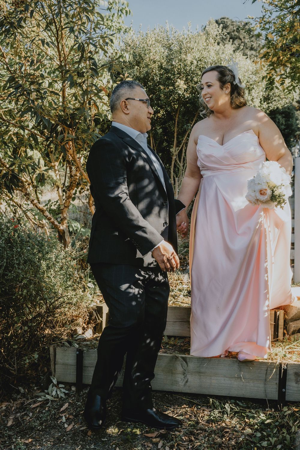 Couple sharing a tender moment in an outdoor garden setting, with one wearing a pink gown and the other in a dark suit.