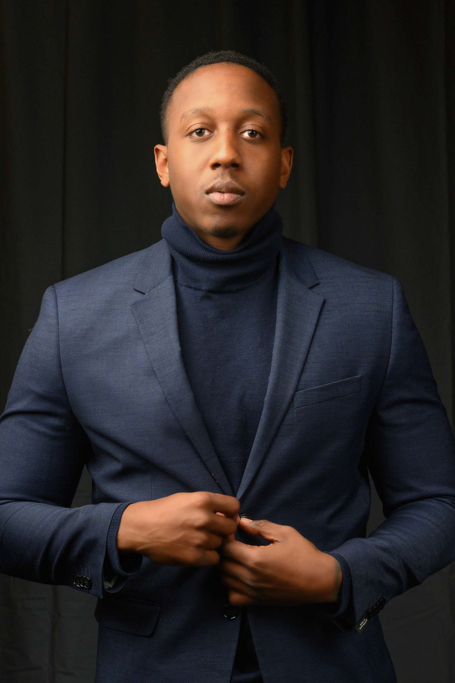 Professional studio headshot portrait of a person wearing a navy blue blazer and turtleneck against a dark background.