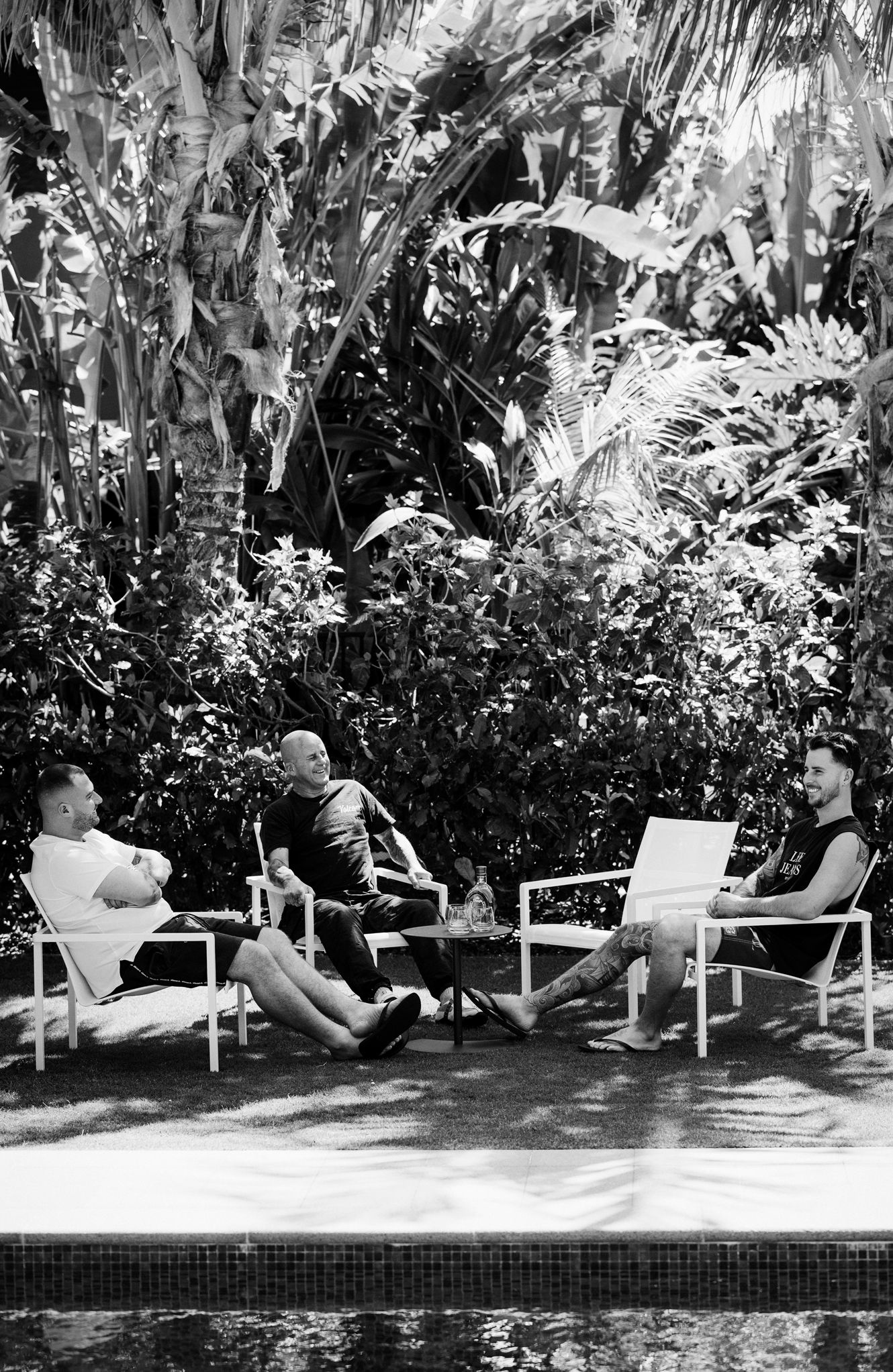 Black and white photo of people relaxing in chairs by a pool surrounded by tropical foliage.