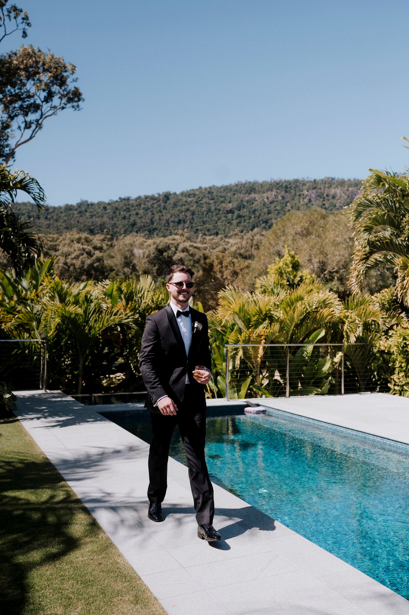 Formal portrait of a person in a tuxedo standing beside a luxurious swimming pool with mountain views.