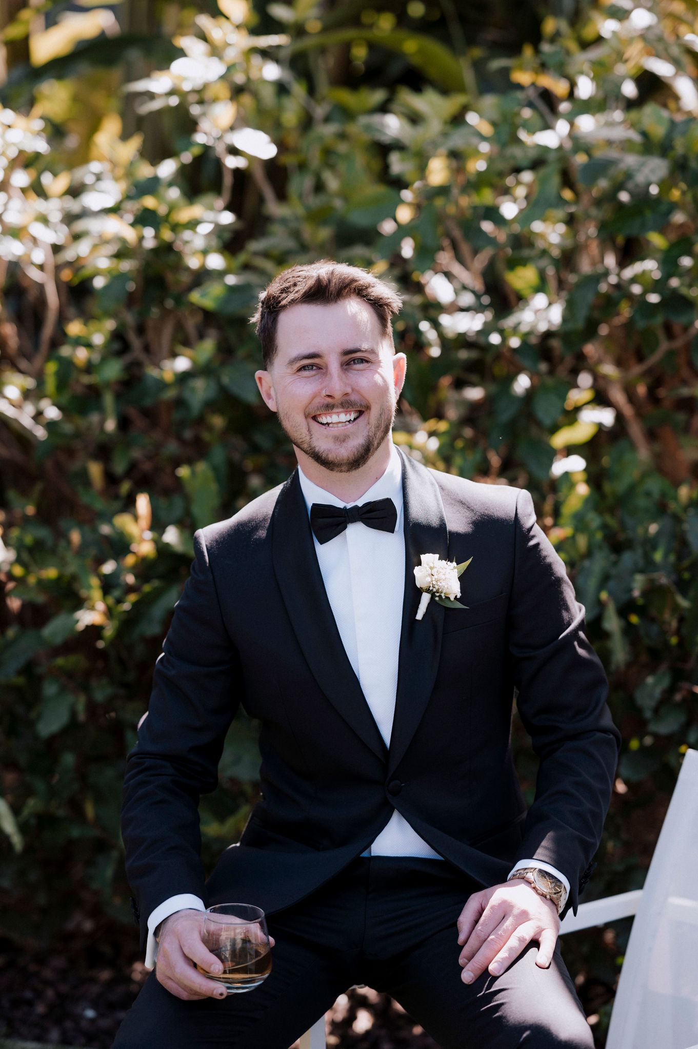 Outdoor portrait of a person in a classic black tuxedo and bowtie against a garden backdrop.