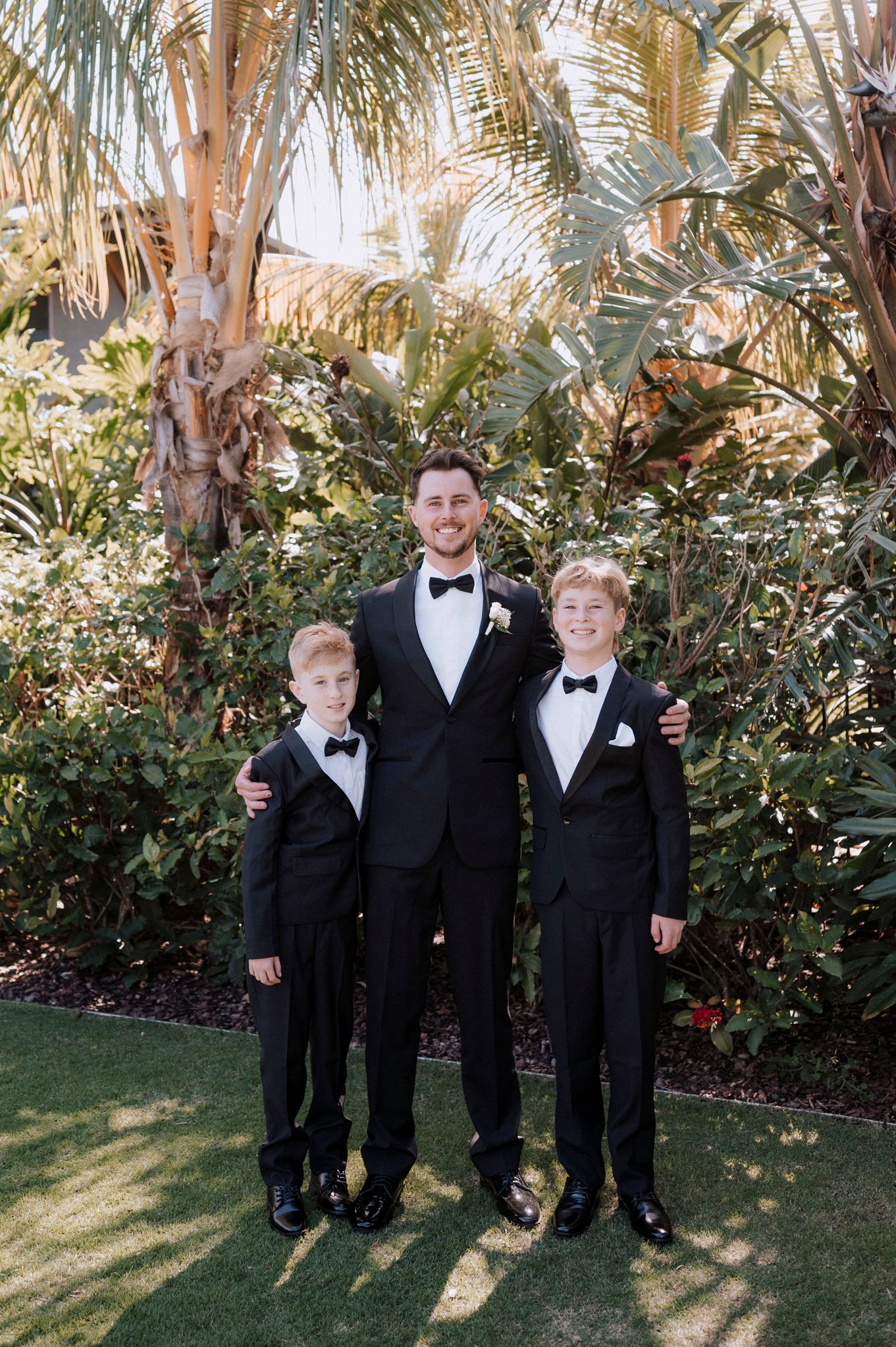 Three people in matching black tuxedos pose together in a lush tropical garden setting.