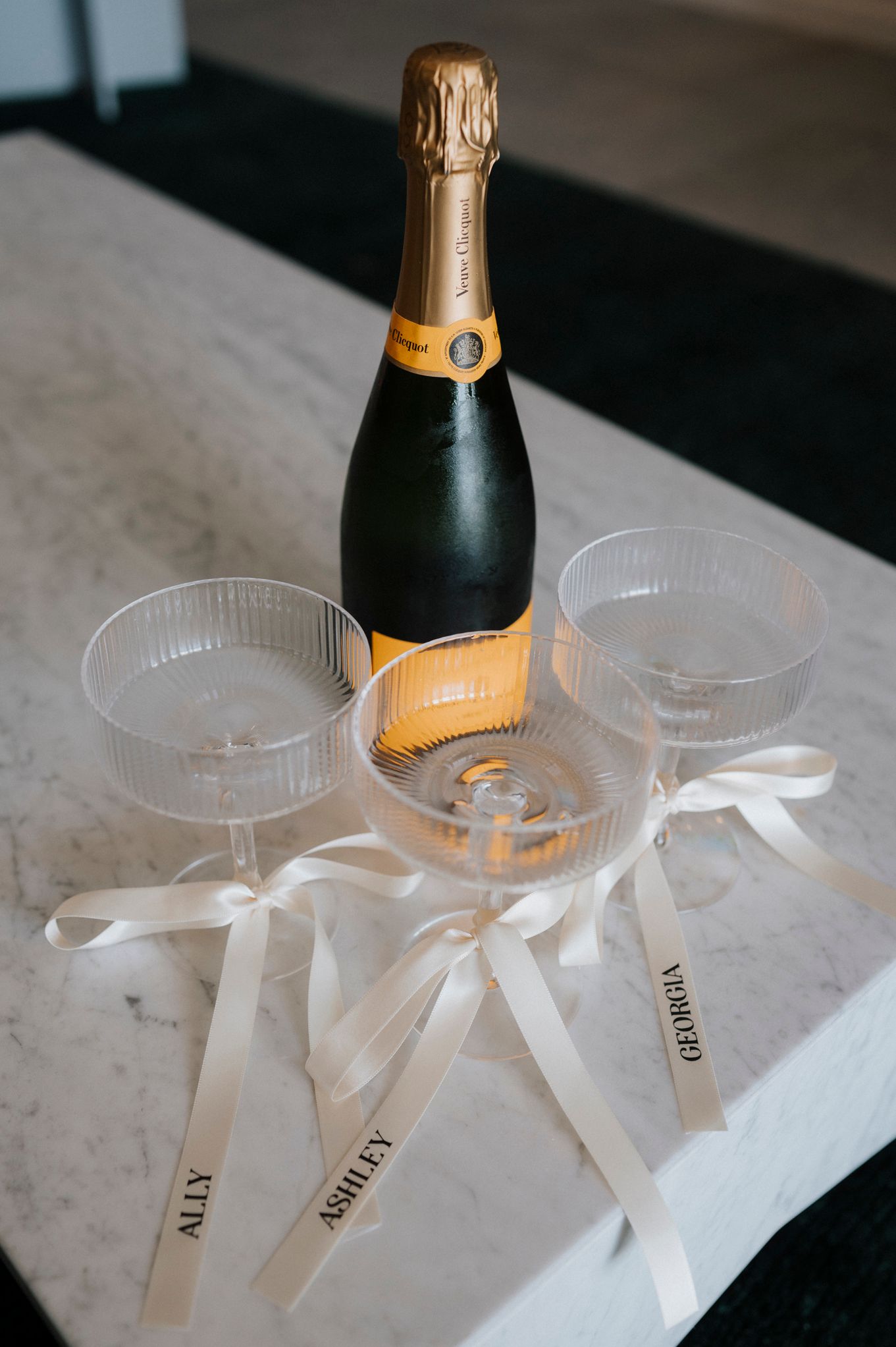 Champagne bottle with three coupe glasses decorated with white ribbons on marble surface.