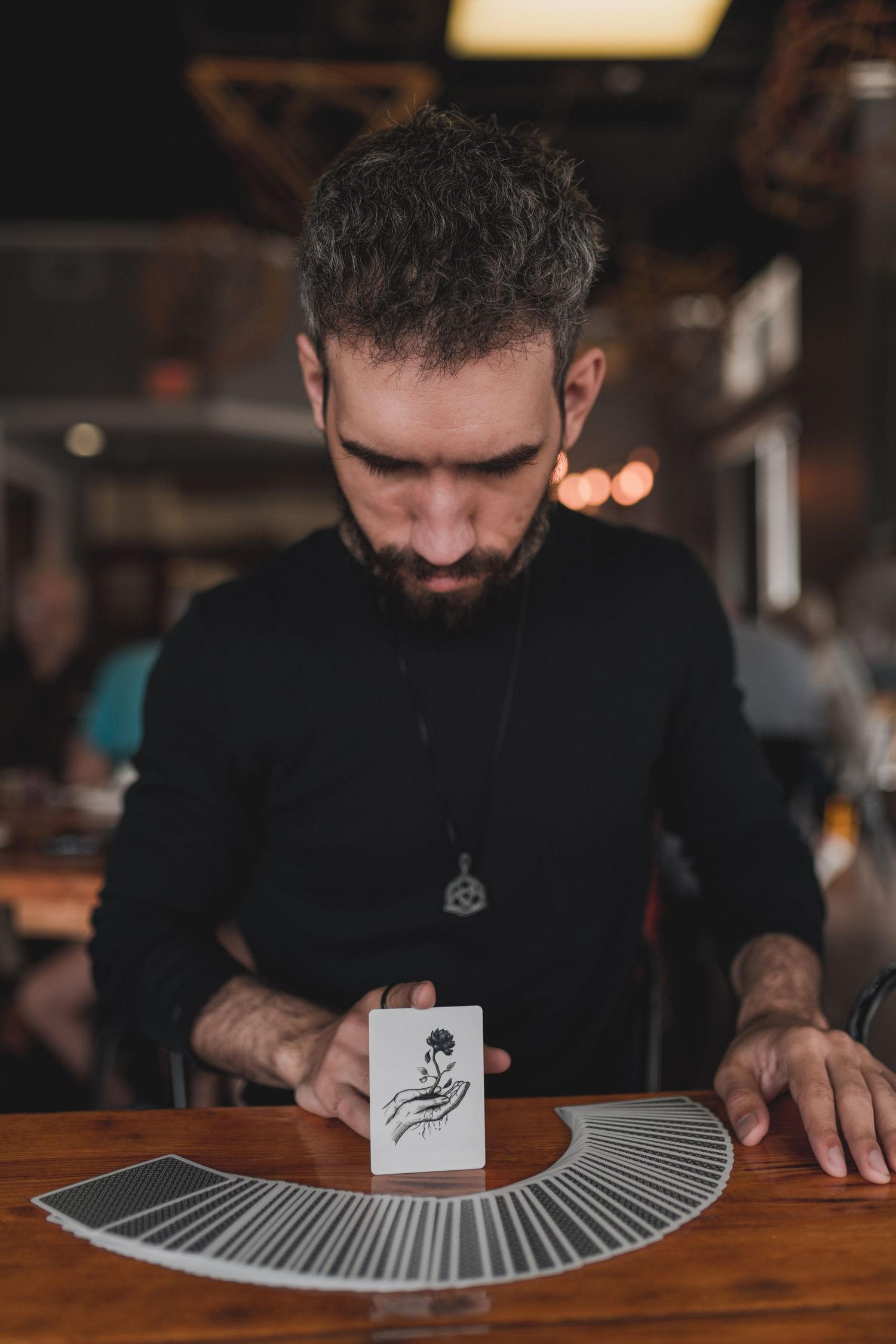 A magician in a black sweater performs a card spread trick on a wooden table in moody lighting.