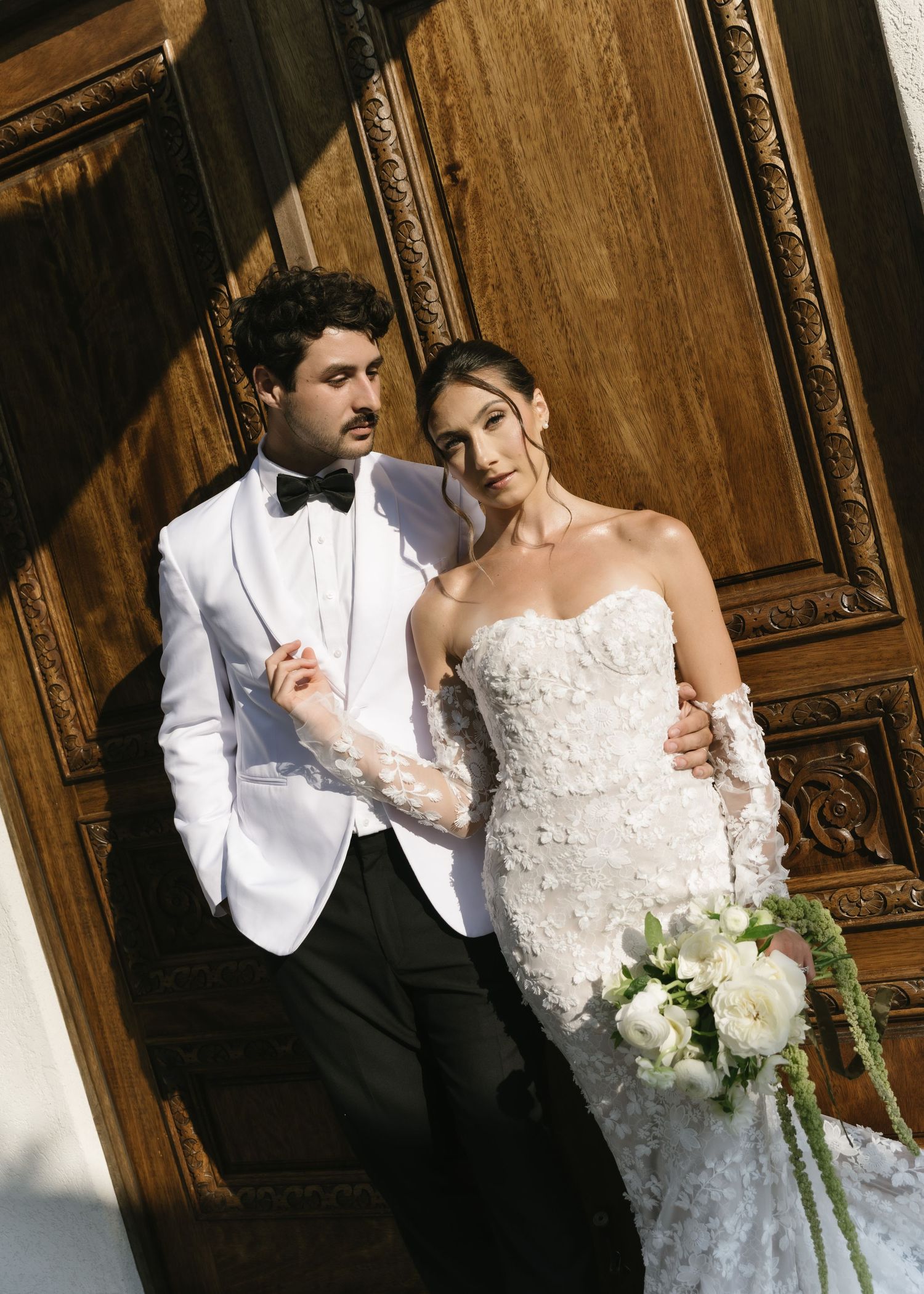 Bride in strapless floral gown holds white bouquet beside groom in white tuxedo jacket, posing before ornate wooden doors.