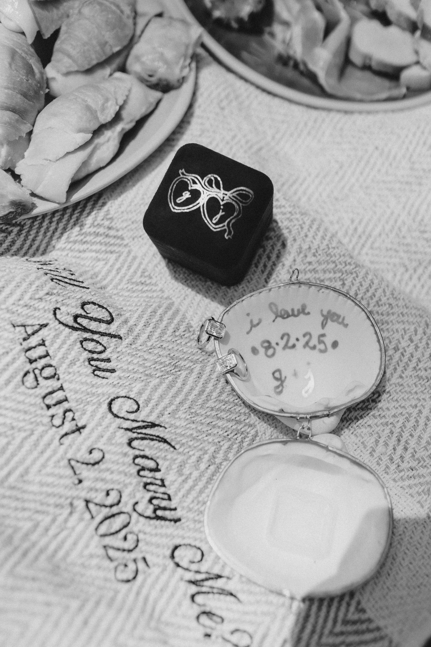 Two engagement rings on top of a seashell ring box on a picnic blanket that reads "will you marry me?"