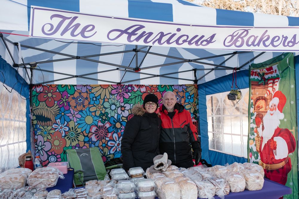 A farmers market booth with The Anxious Bakers banner selling baked goods under a blue canopy tent with colorful wall art.