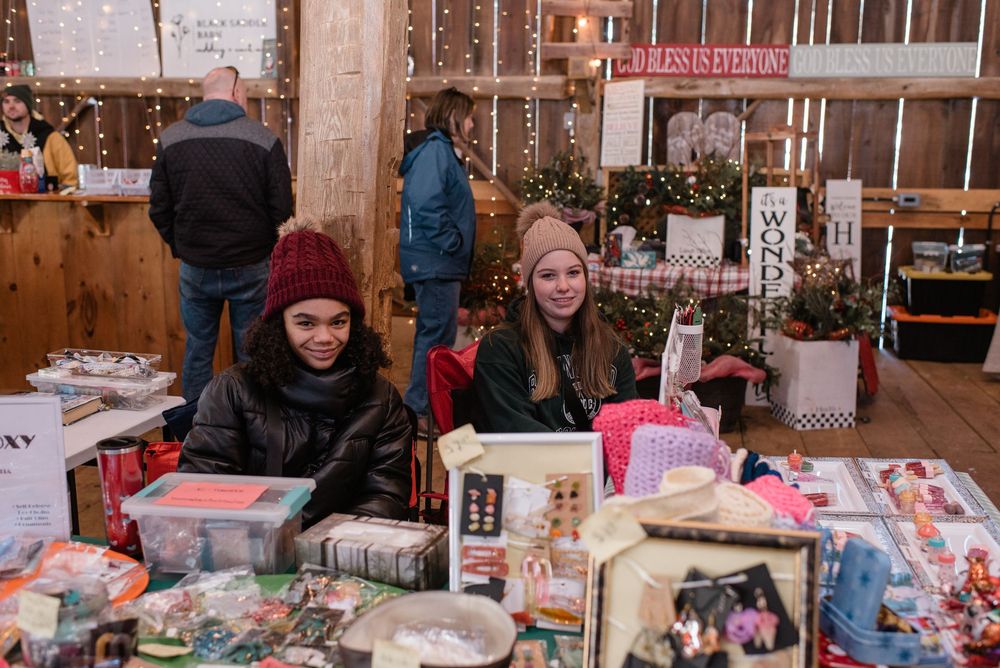 Vendors sitting at a rustic holiday market booth with wrapped gifts and decorations in a wooden barn setting.