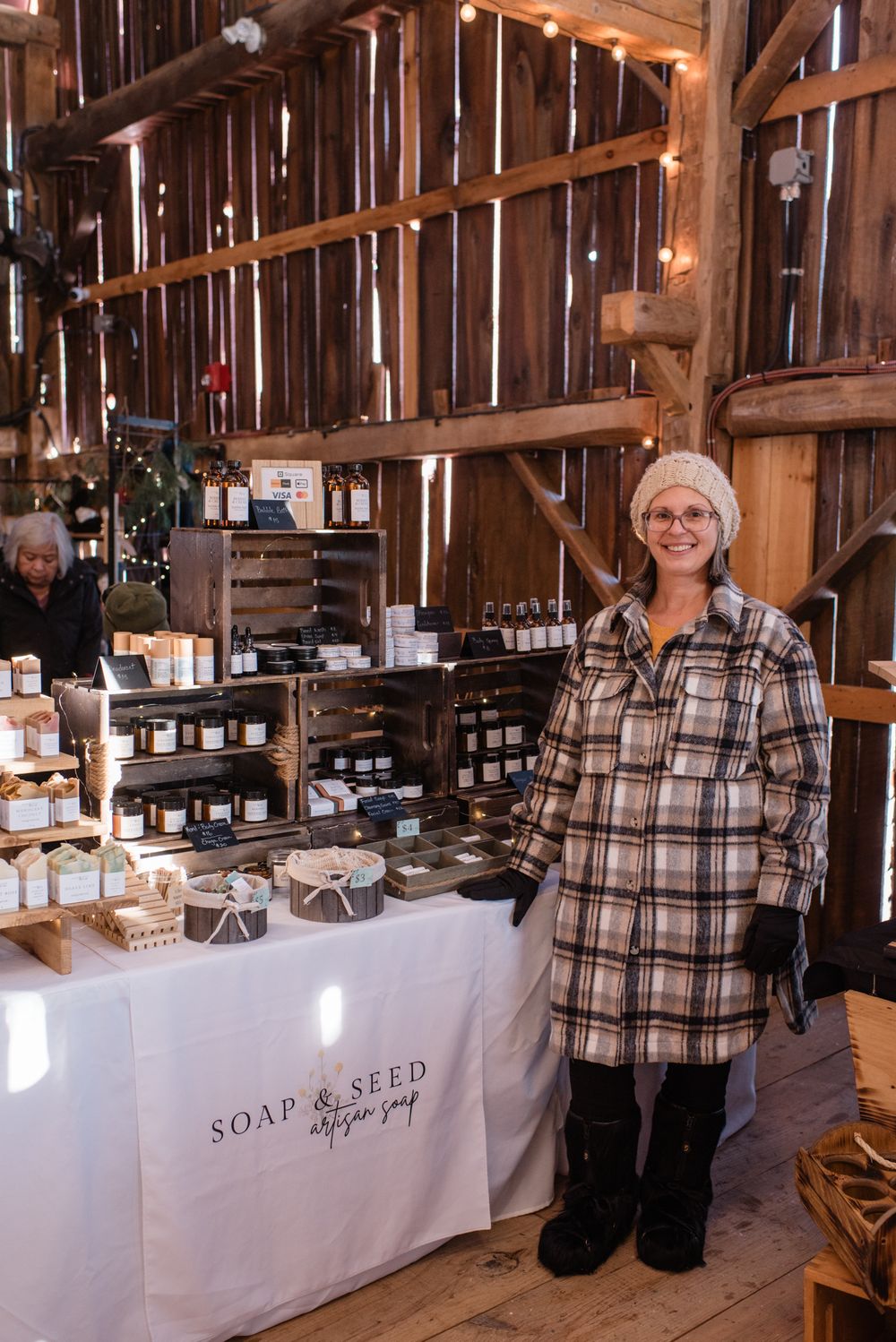 Vendor stands at craft market display table with handmade soaps and products inside rustic wooden barn interior.