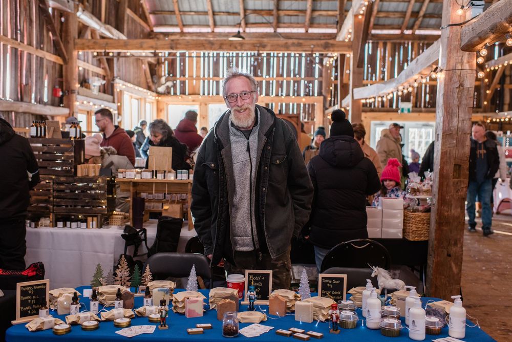A vendor stands behind a table displaying pottery and handmade items at a rustic indoor market.