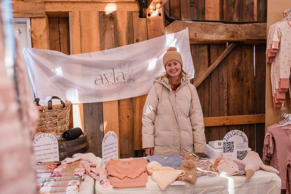 Artisan vendor displays handmade products at craft market booth with banner and winter decor setup in rustic wooden setting.