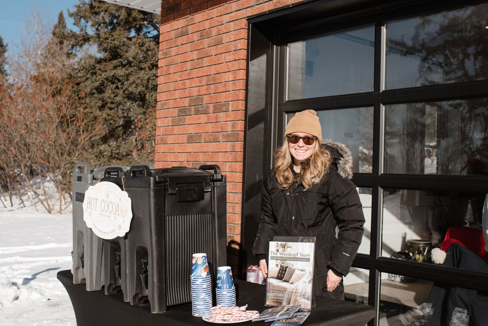 Someone in winter attire stands at an outdoor merchandise display with clothing and drinks on a snowy day beside a brick building.
