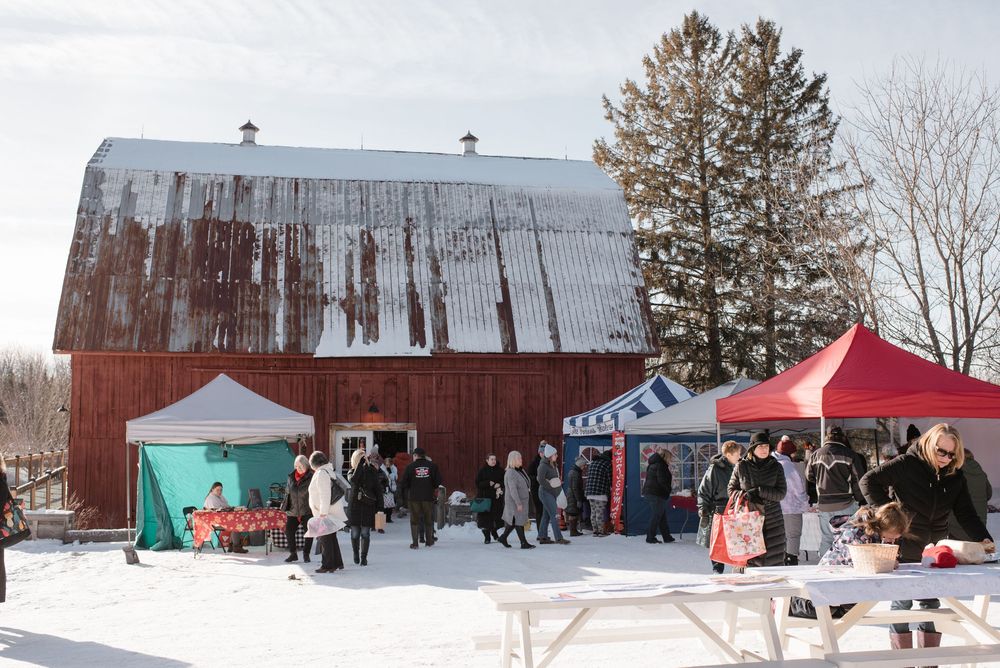 People gather at a winter market with colorful tents outside a large red barn in the snow.