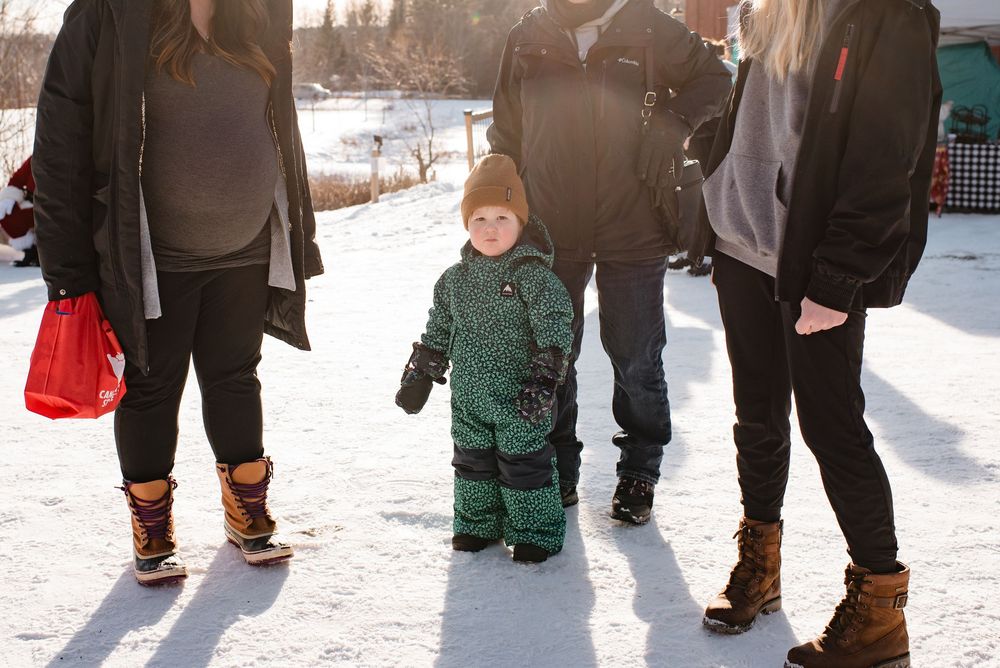 A small child wearing a green snowsuit stands in snow between adults wearing winter boots and warm clothing.