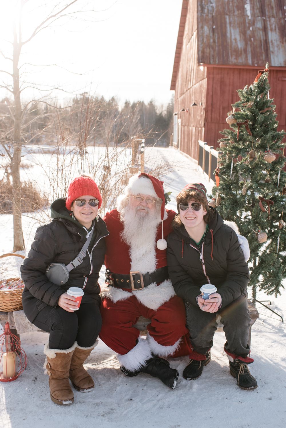 Three people in winter clothing pose with Santa Claus next to a red barn and Christmas tree in snowy scenery.