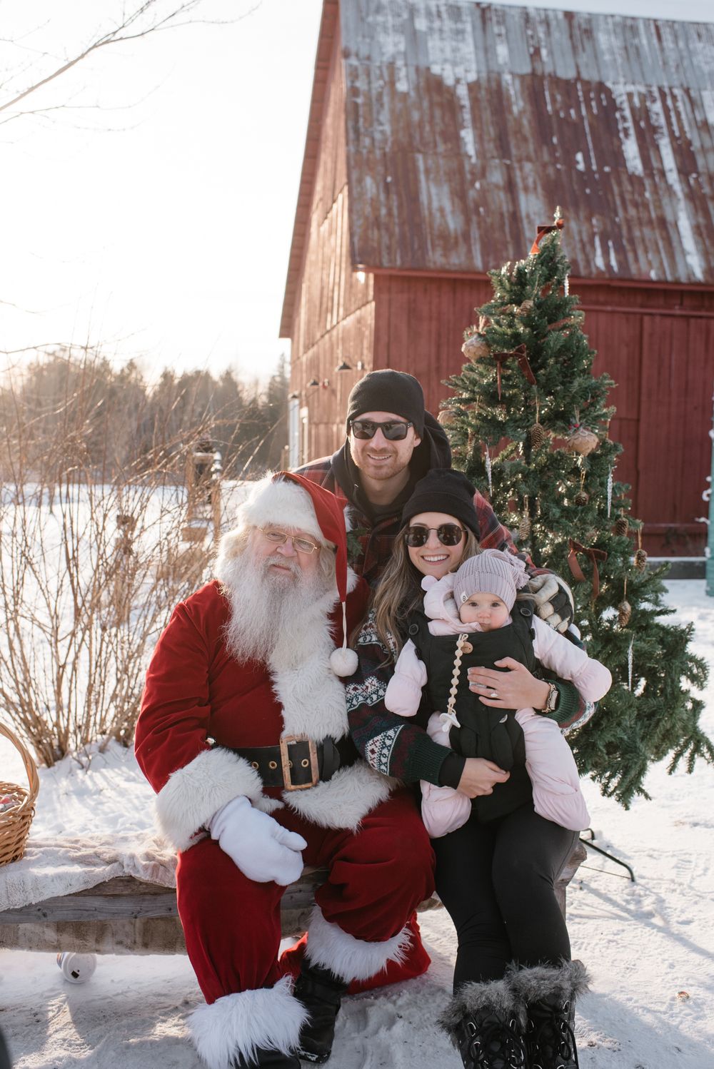 People pose together with Christmas trees in the snow beside a rustic red barn in a series of winter photos.