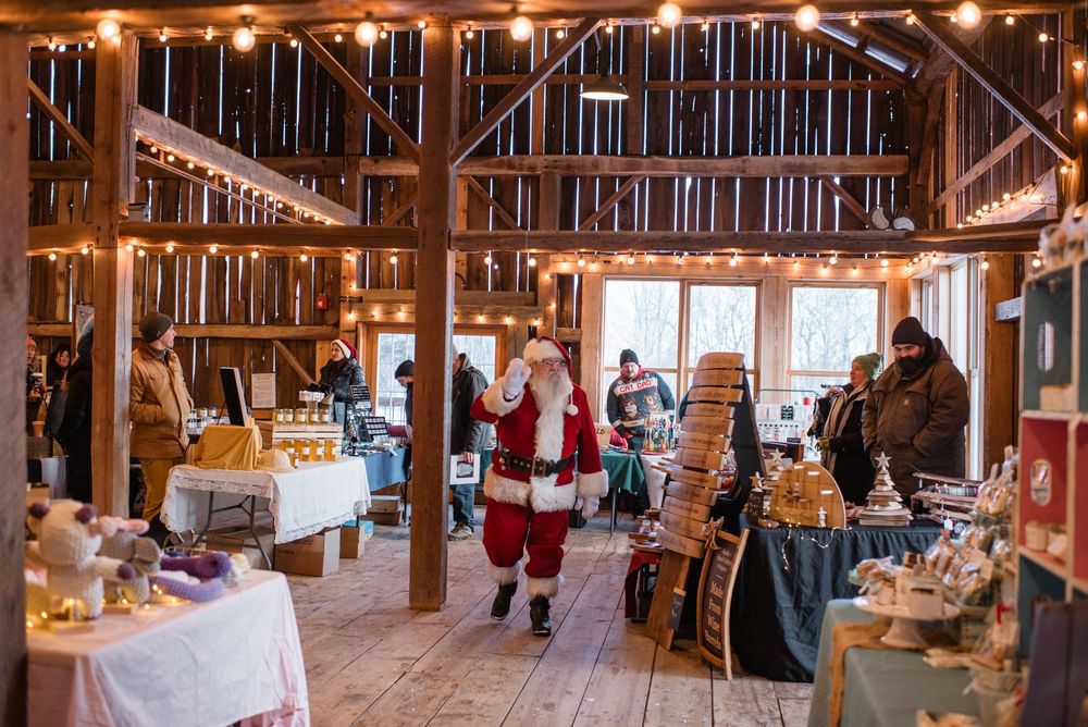 Santa Claus walks through a rustic barn-style market decorated with string lights and vendor tables during holiday season.