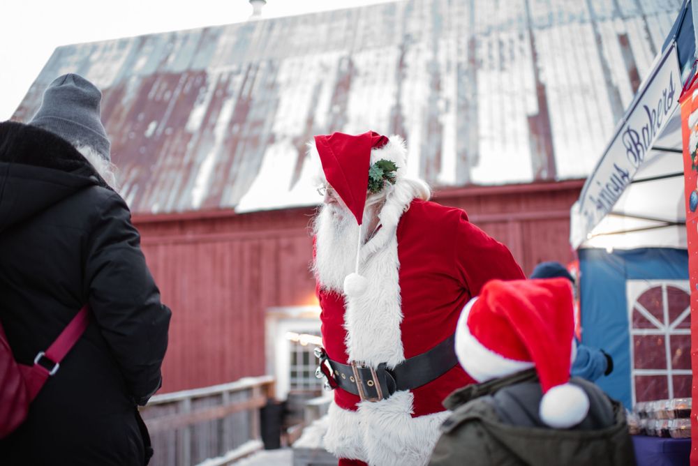 Winter celebration outside a red barn with holiday figures in traditional seasonal outfits playing in the snow.