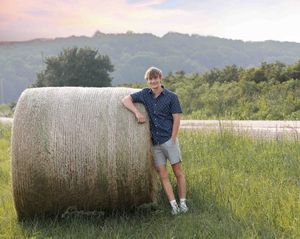 Person posing next to large round hay bales in a grassy field at sunset.