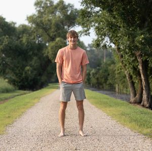 Person wearing a peach shirt and shorts walking on a gravel path through a wooded area.