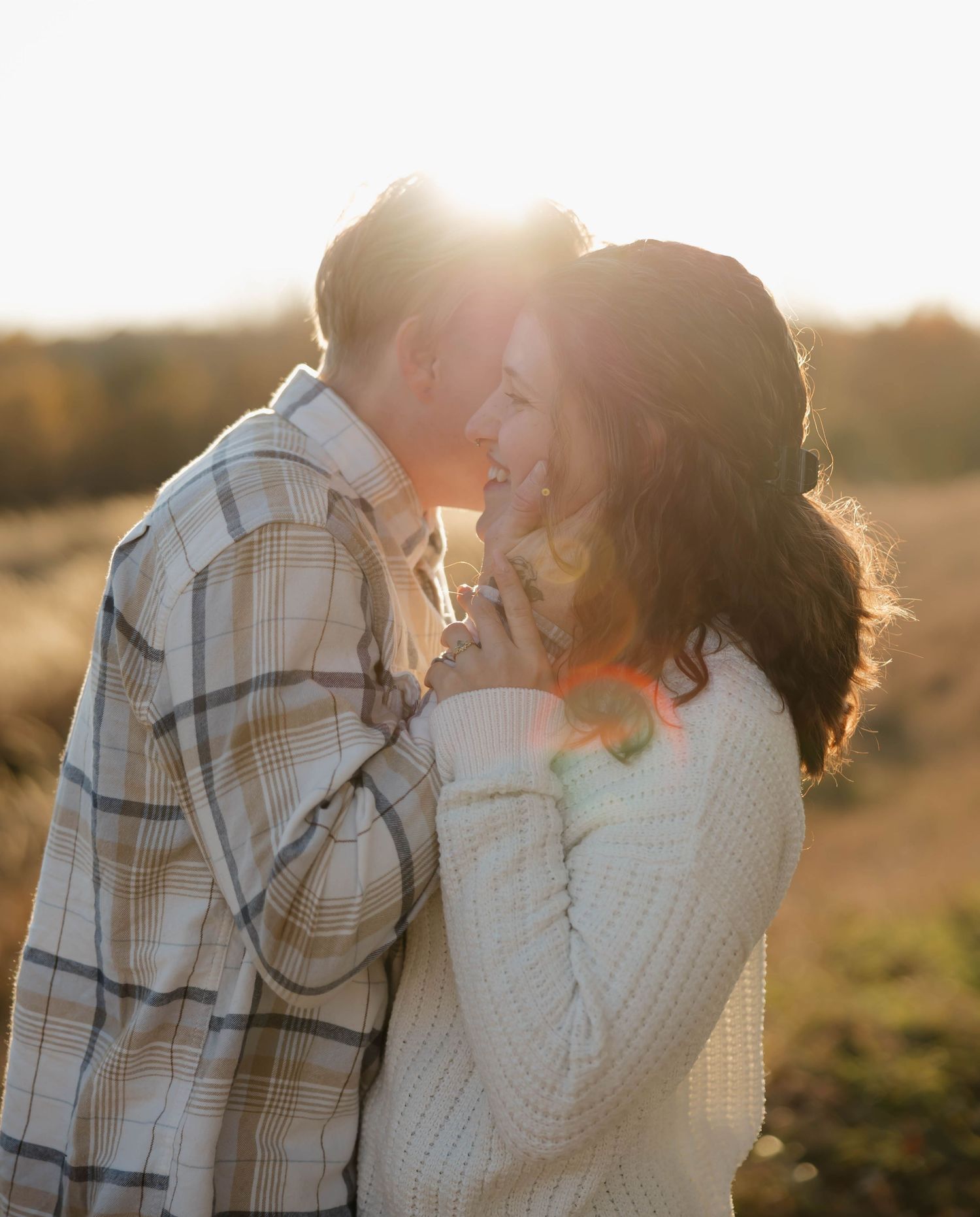 A romantic couple embraces in a field at sunset with golden light streaming through their silhouettes.