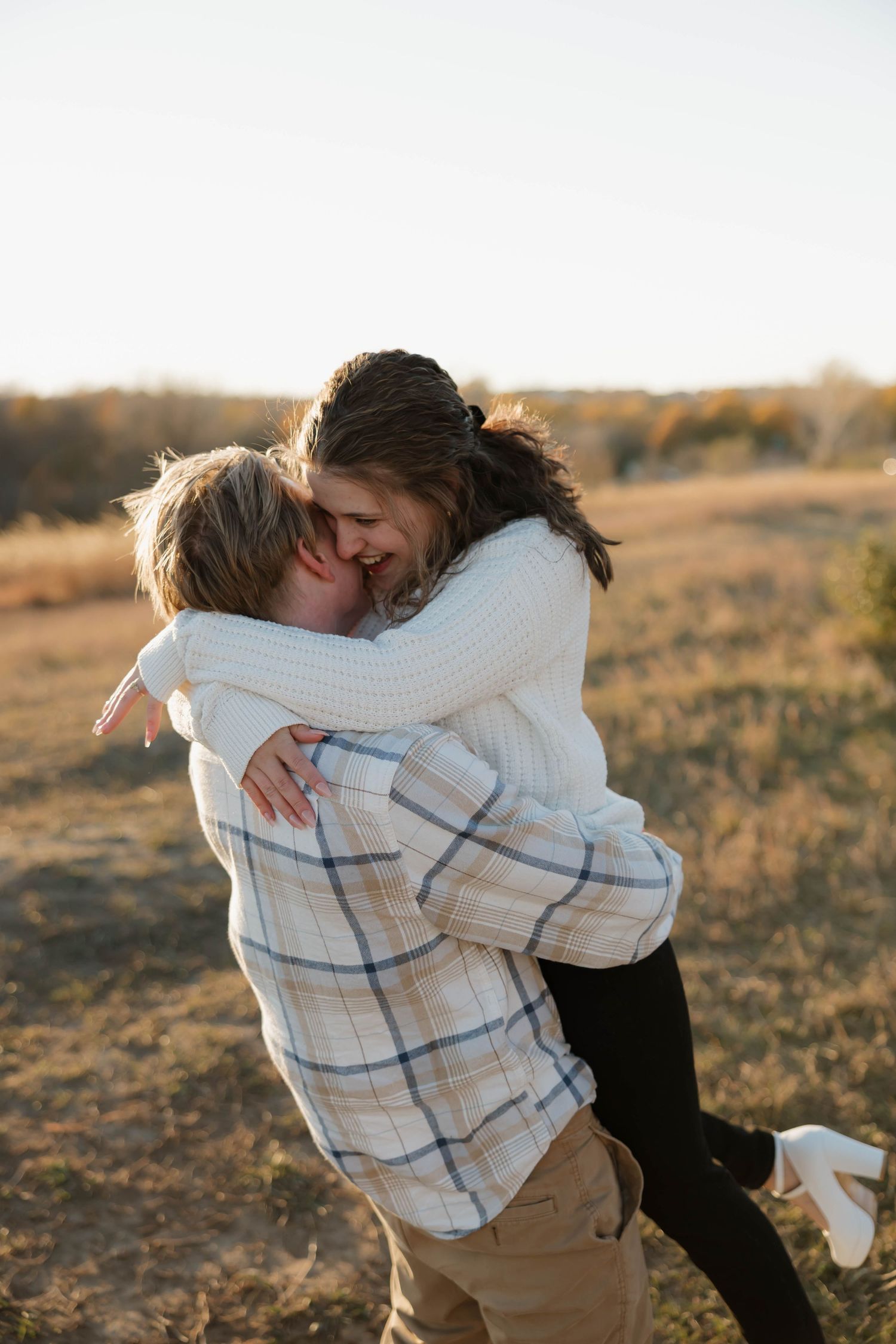 A couple embraces in a warm sunset light on a grassy hillside wearing casual autumn clothing.