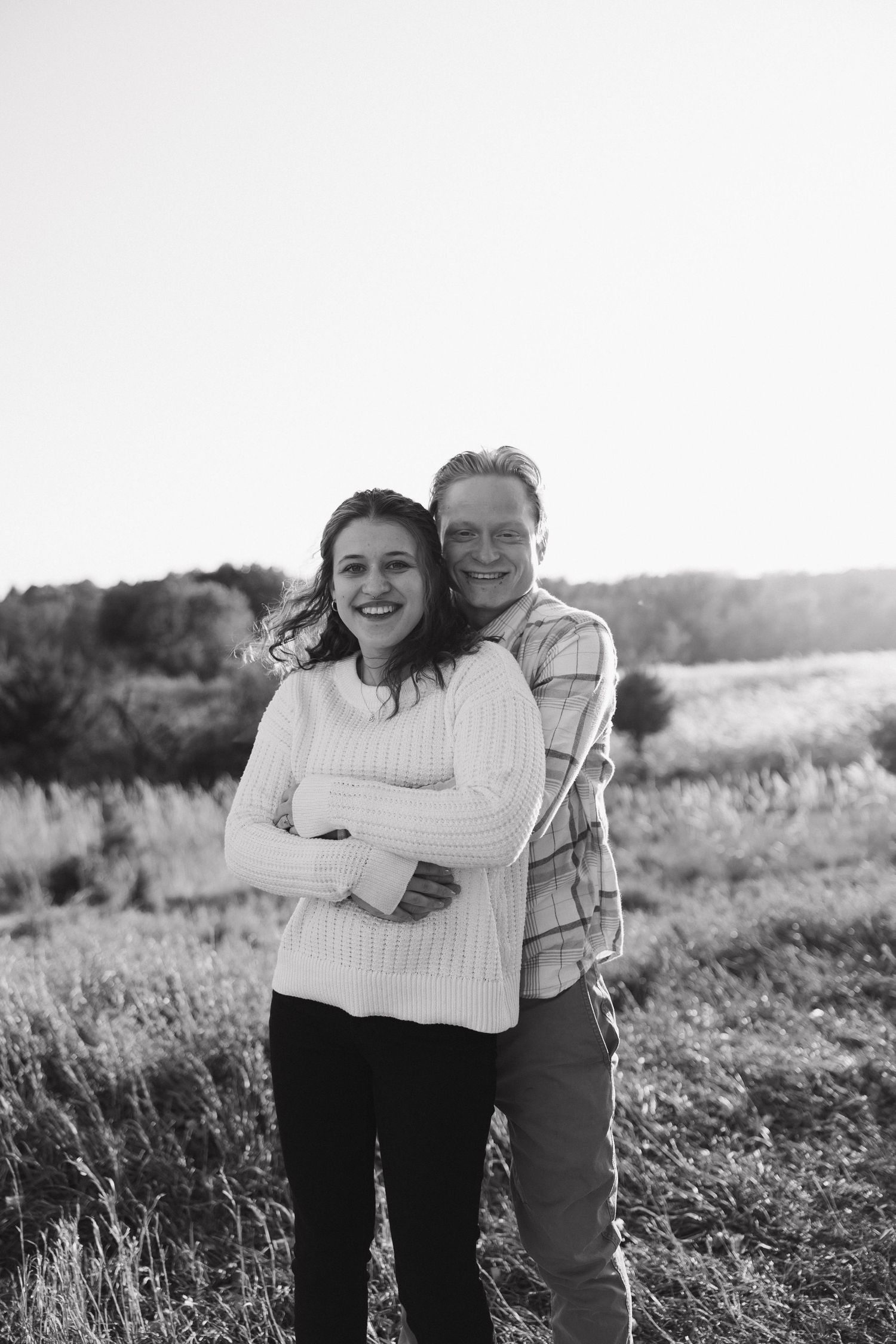 A couple embraces and laughs together in a sunny field with tall grass in black and white photography.